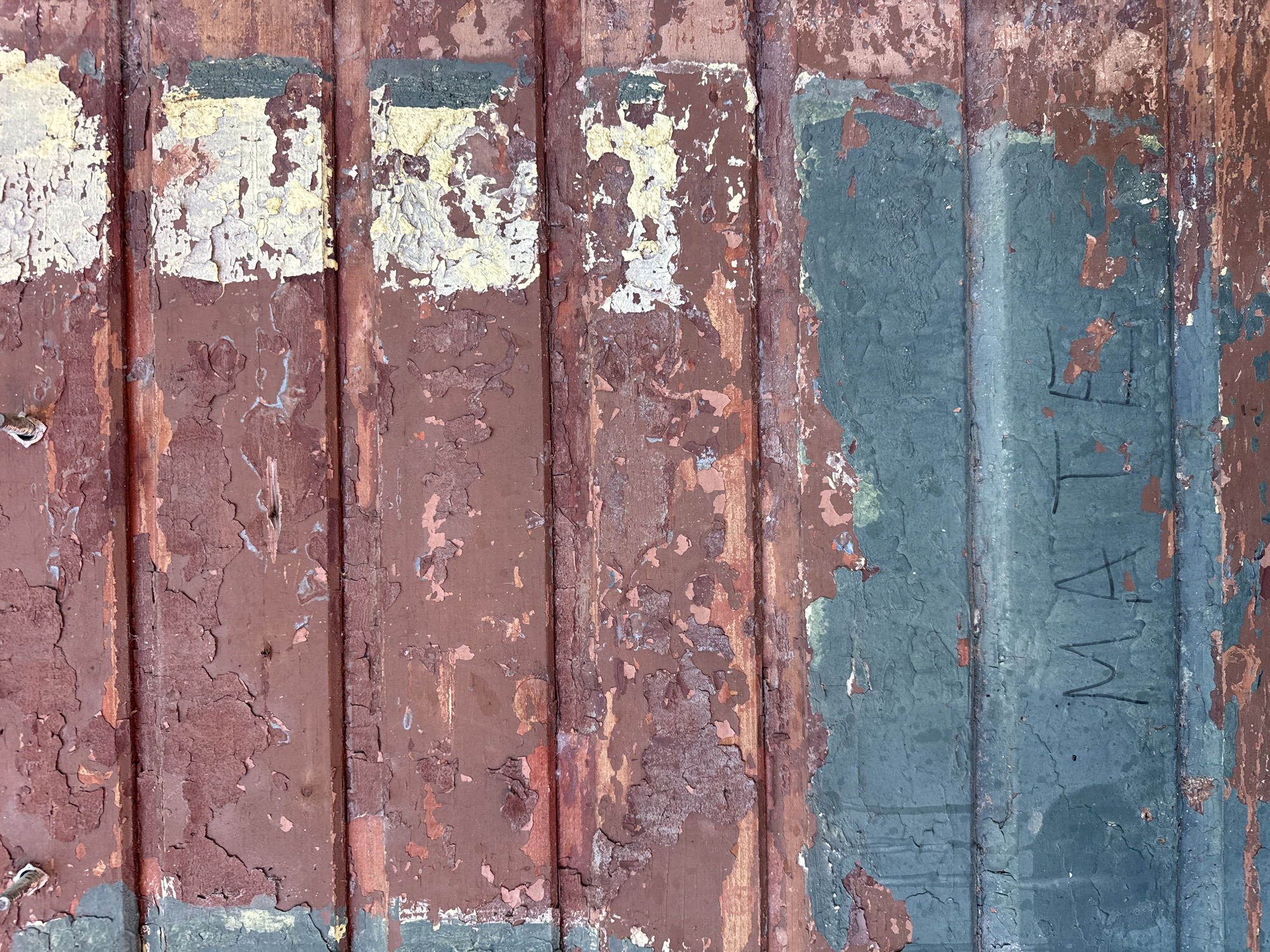 The image depicts a close-up, textured view of weathered wooden planks, likely a wall or fence, in Buknari, Georgia. The wood is covered in peeling paint, revealing layers of red, cream, and a teal or dark green hue. There are also some white and yellow patches where the paint has flaked off significantly. The bottom section of the image features a teal painted surface with the word "MATE" scrawled in black, likely charcoal or a dark marker. The overall impression is one of age and decay, with the vibrant, albeit faded, colors contrasting with the rough, cracked texture of the wood.