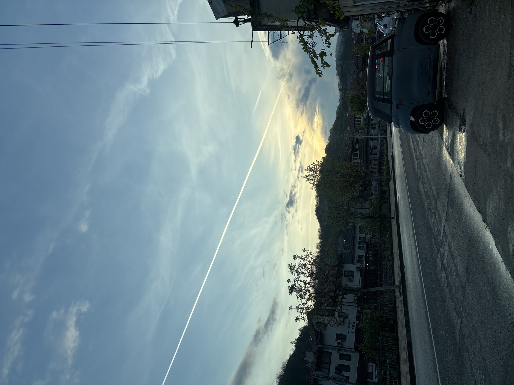The image captures a serene street scene in Zlatograd, Bulgaria, during either sunrise or sunset, marked by a low sun casting a warm, yellowish glow on the horizon. The sky, occupying the upper two-thirds of the frame, displays a mix of clear blue and scattered, wispy clouds. A prominent white airplane contrail diagonally streaks across the sky from the upper left. Thin black utility lines are visible against the upper left sky.
In the mid-ground, tree-covered hills or mountains form a natural backdrop. Below, several buildings, some featuring traditional architecture, line the street. On a white building on the left, partially visible text reads "КЪЩА ЗА ГОСТИ" (Guest House) and "ТОНИ" (Toni). Bare trees and some leafy branches are scattered throughout the scene, especially near the buildings and along the hills.
The asphalt road occupies the bottom portion of the image, appearing damp or wet in places, reflecting the ambient light. A compact, grey-blue car, possibly a Citroën Ami, with distinctive white and black hexagonal wheel covers, is parked on the right side of the road, facing away from the viewer. The overall atmosphere is quiet and tranquil, depicting a peaceful moment in the town.