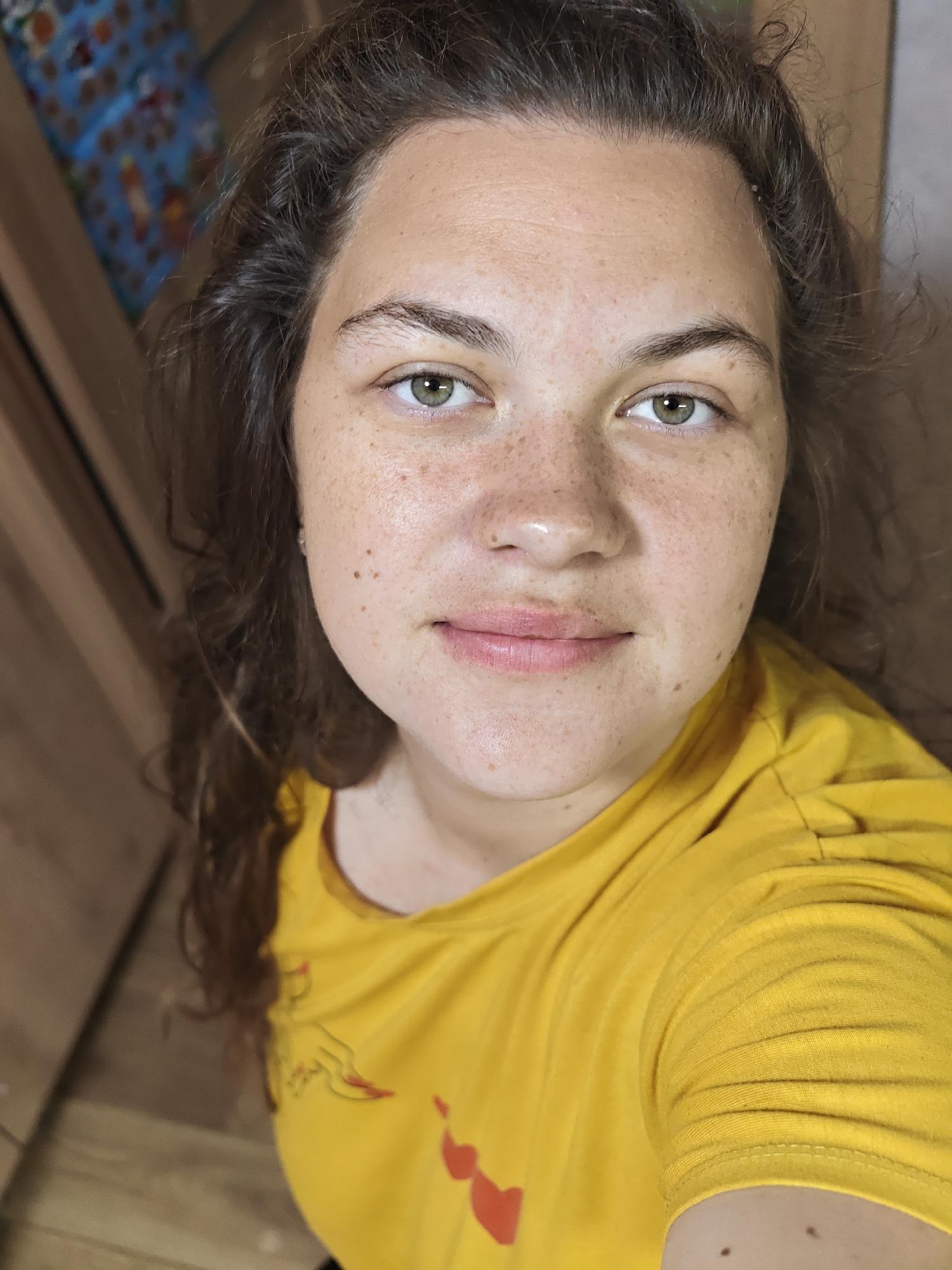 A close-up, eye-level selfie captures an adult female looking directly into the camera. She has long, dark brown, wavy hair styled away from her face, revealing numerous light brown freckles across her forehead, nose, and cheeks. Her eyes are light green or hazel, with visible pupils and distinct catchlights. Her eyebrows are naturally shaped and dark. Her lips are closed in a neutral or faint, slight smile. She is wearing a bright yellow, short-sleeved t-shirt with subtle red graphic elements, appearing to be small hearts and possibly a stylized hand, on the left chest area. The indoor background is significantly blurred, featuring warm-toned wooden surfaces on the left side and a less distinct, lighter-colored wall or surface on the right. In the far upper-left corner, a highly blurred object with dominant blue and orange patterns is visible. The image does not contain specific visual information to determine a precise geographical location, such as Chaikovsky, Russia.