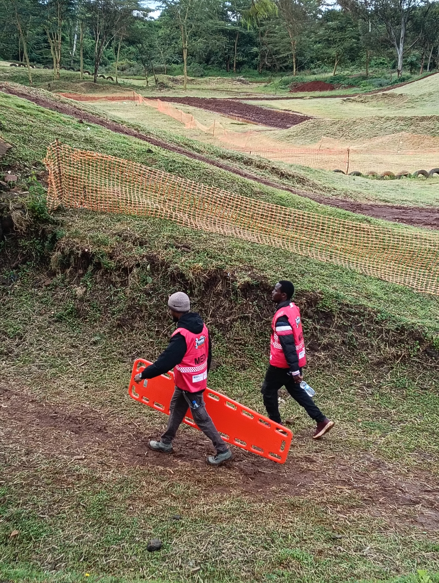 The photograph depicts a rural area on the outskirts of Nairobi, Kenya. In the foreground, two individuals wearing red vests marked "MEDIC" carry an orange stretcher across a grassy, muddy terrain. The surrounding environment consists of a grassy hillside with a safety fence and a dirt race track visible in the background. Tires line the track. The weather appears overcast.