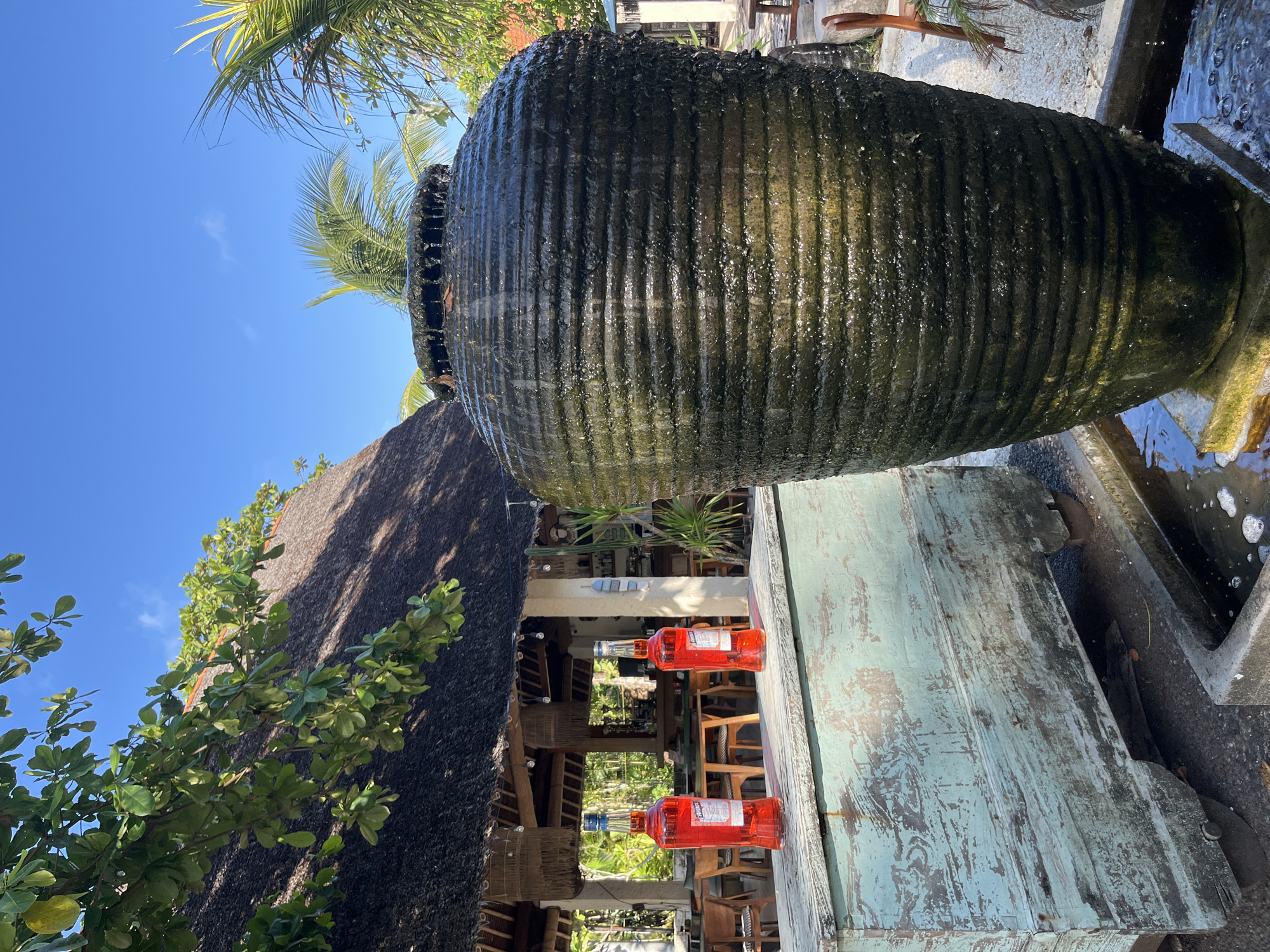 A large, dark green, textured ceramic urn fountain with water cascading down its sides is the primary subject of the image. To the left of the fountain, on a distressed, light blue wooden chest on wheels, sit two identical bottles of red liquid, possibly wine or liqueur. Behind these objects, a thatched-roof building with a rustic, open-air dining area is visible, with tables and chairs set up. Palm trees and lush green foliage are interspersed throughout the scene, suggesting a tropical outdoor setting. The sky is clear and blue. The image appears to be taken in Kuta Selatan, Indonesia, likely at a restaurant or bar.
