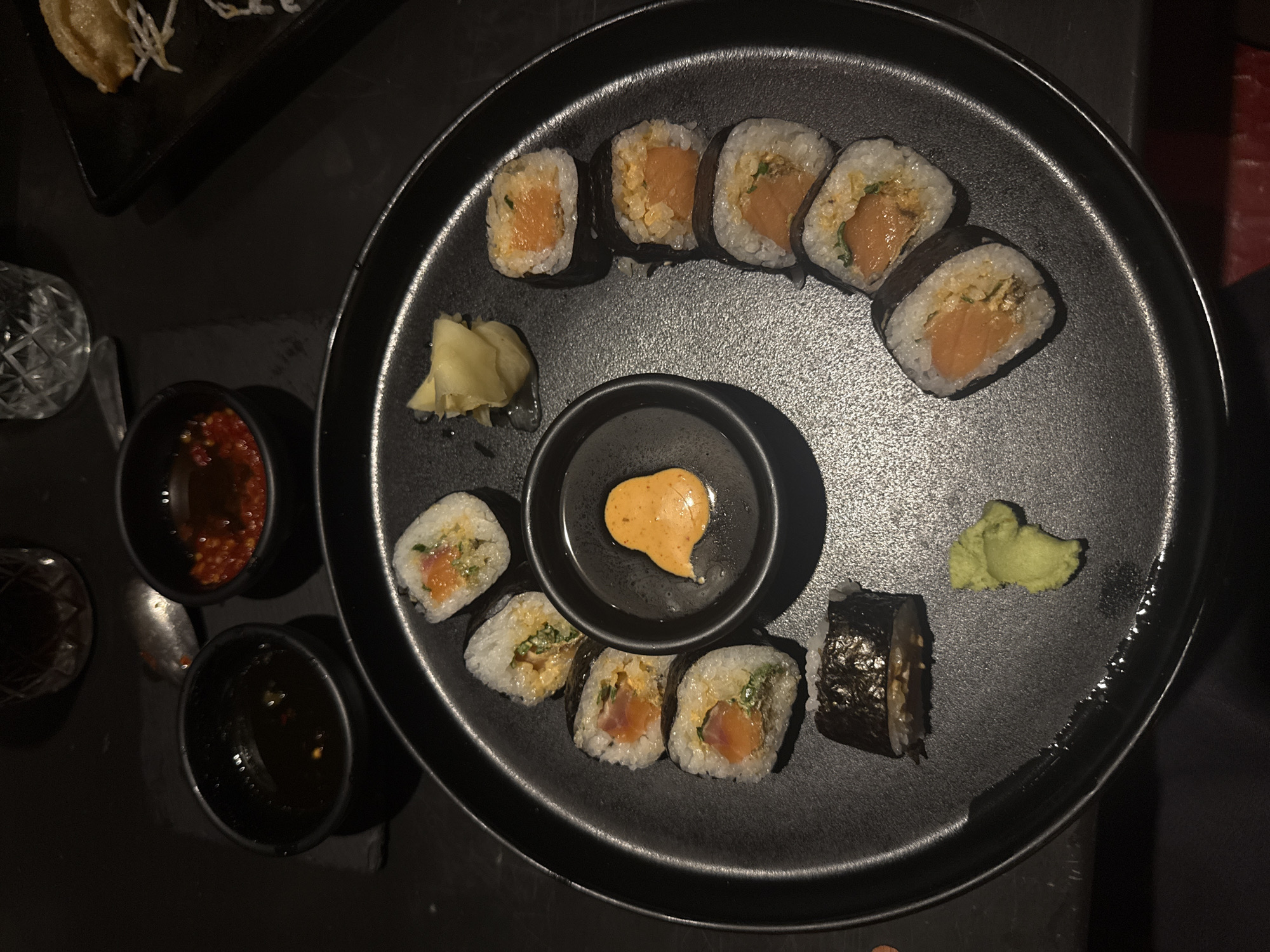 The image depicts a black plate of sushi, featuring various rolls arranged around a smaller bowl of orange sauce, with a dollop of wasabi and a pile of pickled ginger. The sushi rolls are filled with salmon and seaweed, and the dish appears to be set in a dim environment. In the background, there are two black bowls containing dark-colored liquids, along with a glass object and part of a black plate with what appears to be a dumpling. The setting is possibly a restaurant in Mexico City, Mexico, based on the presumed food and service style. The image shows a static arrangement of food on a table.
