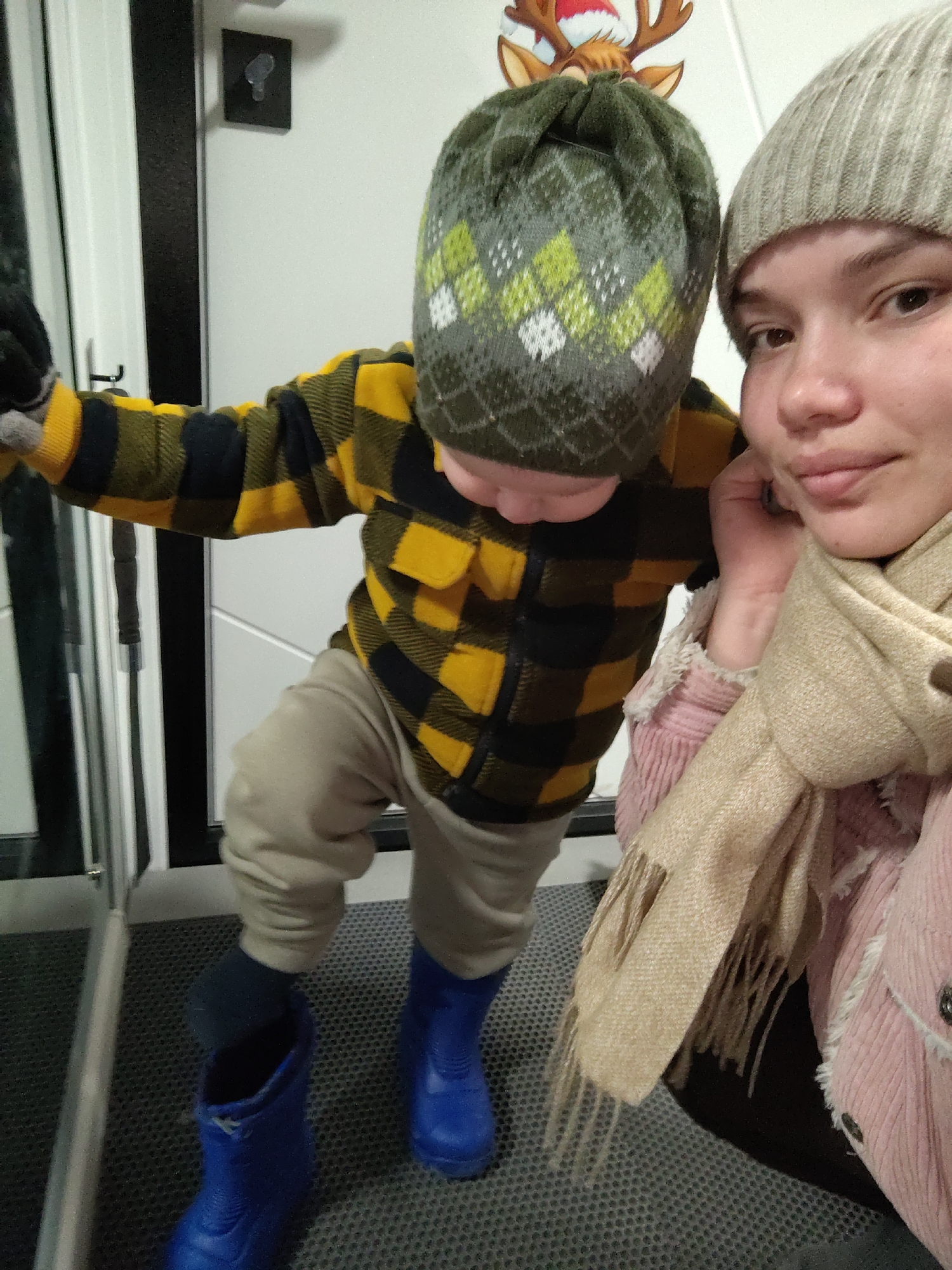 A woman and a young child are captured in an indoor selfie, likely in an entryway. The woman, positioned on the right, wears a light grey knit beanie and a beige scarf over a pink corduroy jacket. She has a neutral to slightly smiling expression. The child, a young boy, is seen leaning forward on the left, clad in a green and white patterned knit beanie, a yellow and black plaid shirt, light tan pants, and bright blue rubber boots. He appears to be in the process of putting on or taking off his boots, with one foot already inside a boot and the other foot, covered by a dark sock, hovering above the second boot.
In the background, a white door frame is visible on the left, and a white wall behind the child features a dark-colored mounted object. Above the child's head, a Christmas-themed reindeer decoration is affixed to the wall. The subjects are standing on a dark grey, textured floor mat, typical of an entrance area. The warm clothing, including beanies, scarves, and rubber boots, suggests cold weather, consistent with the provided location context of Марковский, Russia, possibly during autumn or winter. No visible text is present in the image.