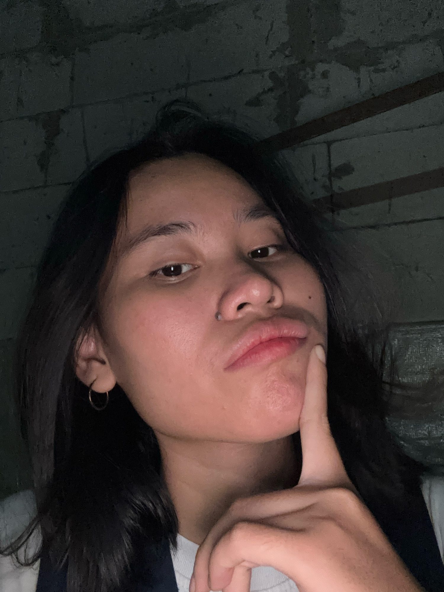 A young person, with dark hair and light brown skin, is shown in a close-up selfie. They are wearing a white t-shirt. Their right hand is raised, with the index finger touching their chin. They have a hoop earring in their left ear and a small piercing above their upper lip. The background appears to be a brick wall with a rough, textured surface, suggesting an indoor or covered outdoor setting. The lighting is dim, casting shadows and highlighting the subject's face. The image quality is moderate, with some graininess. No specific location in East Jakarta, Indonesia can be determined from the visual information provided.
