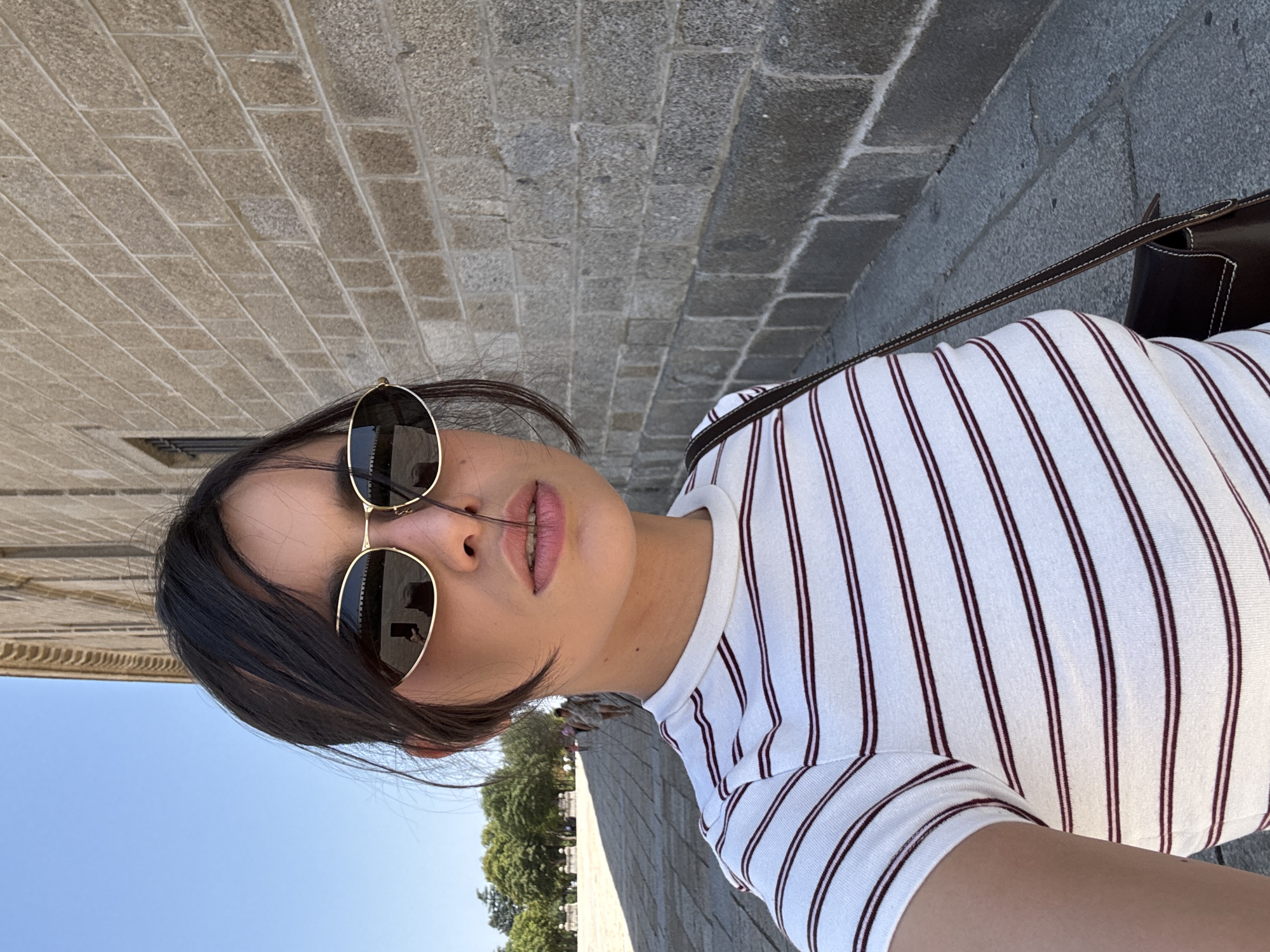 The image shows a woman taking a selfie outdoors on a sunny day in the city of San Lorenzo de El Escorial, Spain. She is wearing a white t-shirt with thin dark red and pink stripes, sunglasses with gold frames, and a dark brown shoulder bag. Behind her, a textured stone wall of a building is visible. To the left, a paved walkway and trees can be seen, with several people walking in the background. The sky is clear and blue.