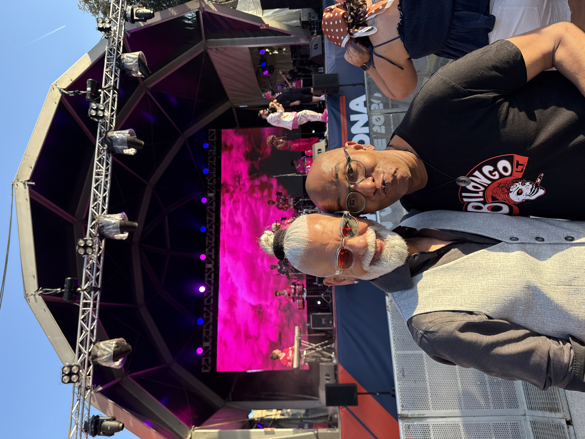 Two men are posing for a selfie in front of a concert stage set up outdoors. The man on the left has white hair styled in a bun, a white beard, and is wearing sunglasses and a grey vest over a dark shirt. The man on the right has glasses, is wearing a black t-shirt with a red graphic, and is making a kissing face. In the background, a band is performing on stage, with musicians playing instruments and a large pink screen displaying abstract imagery. The stage is equipped with numerous lights and speakers. The event appears to be taking place during the day, as indicated by the blue sky visible above the stage structure. The text "ONA 2023" is visible on a barrier in the foreground, suggesting the event might be related to a festival or organization in Barcelona, Spain.