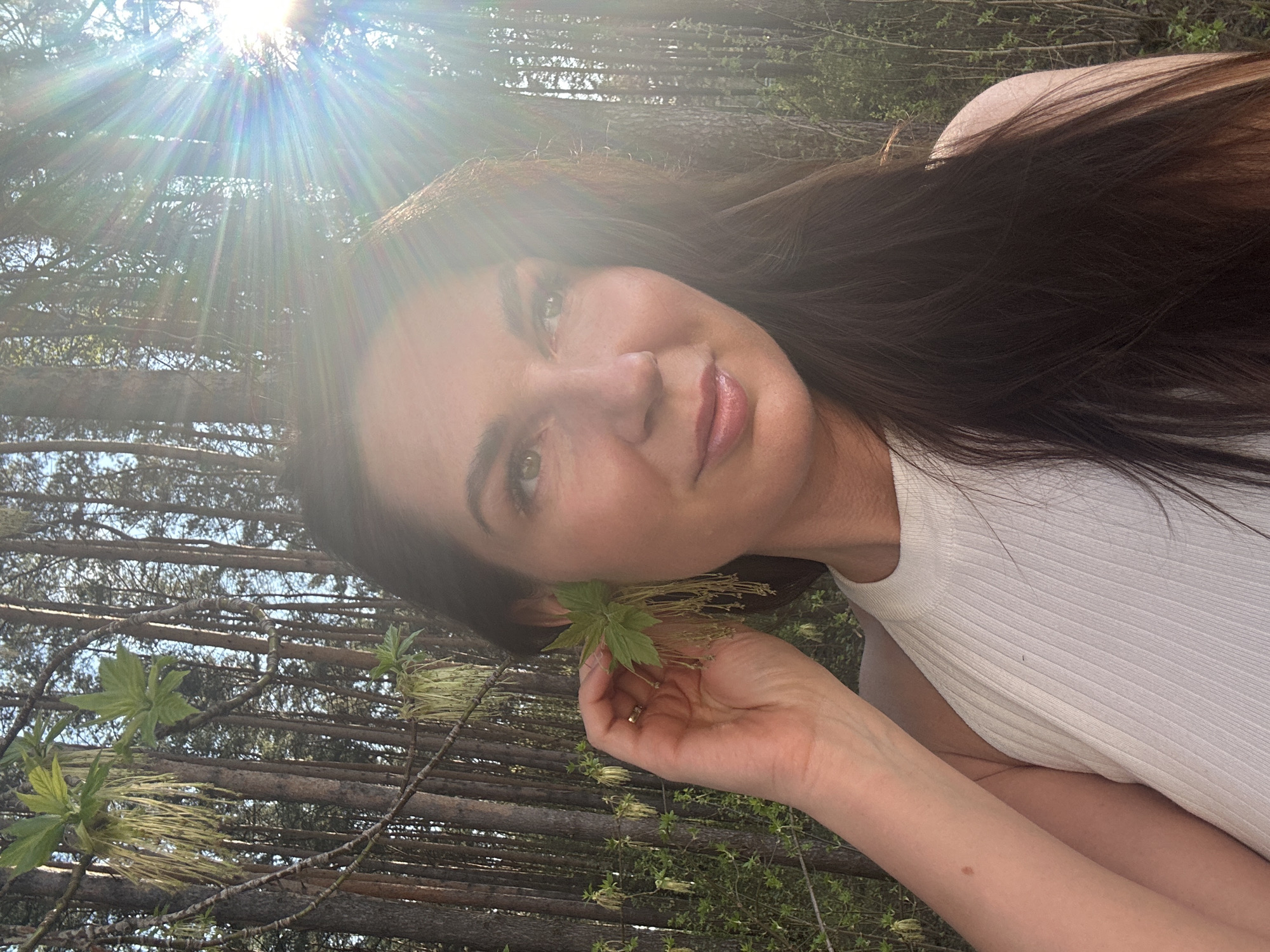 A woman with long dark hair is holding a green leaf with yellow tassels in her right hand. She is wearing a white ribbed top. The background consists of trees with thin trunks and branches. The image is taken outside in daylight, with bright sunlight filtering through the trees. The location is Chaikovsky, Russia.