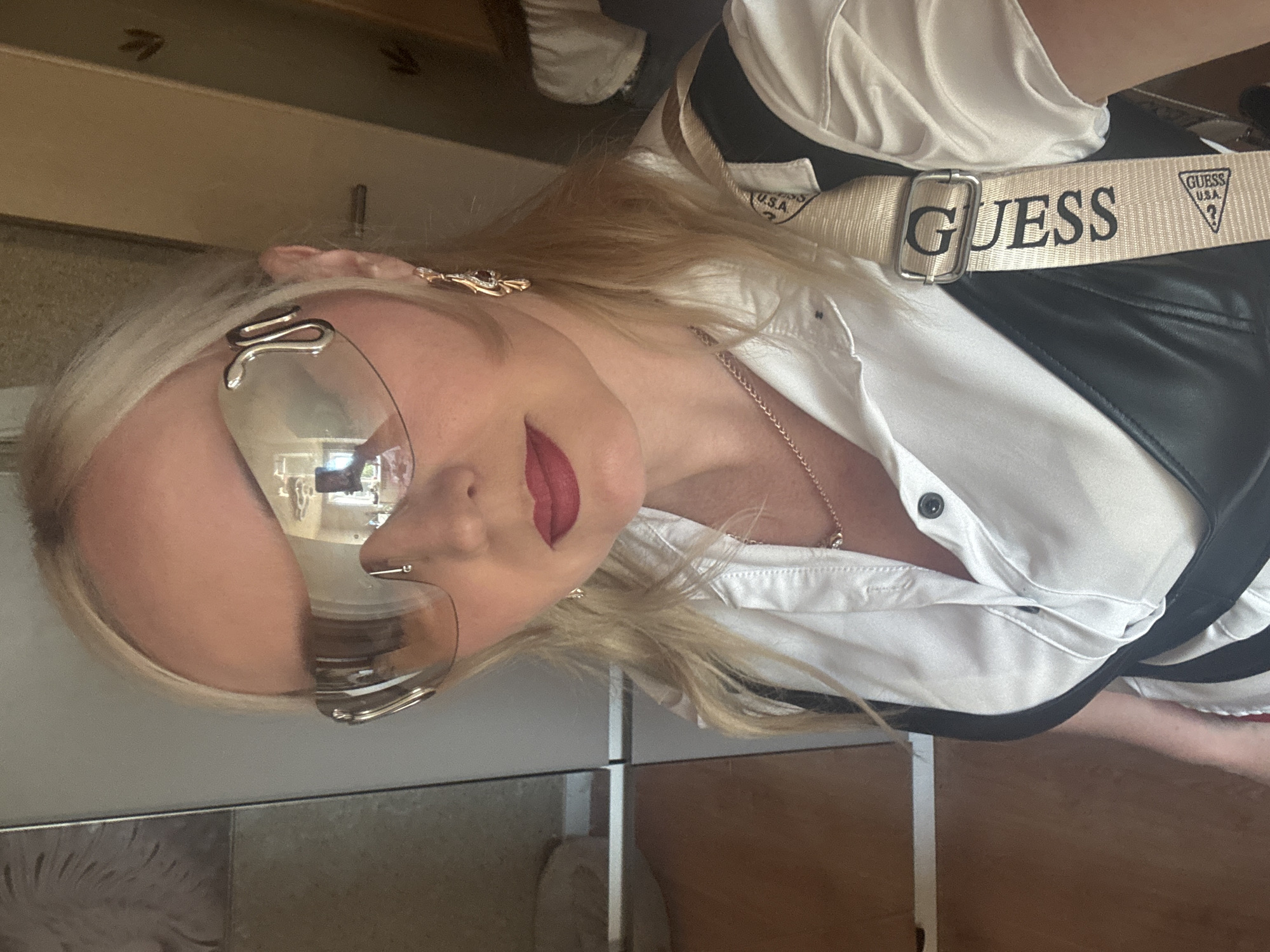 A blonde woman wearing large, futuristic sunglasses and a white satin shirt with a black bustier is taking a selfie. She has on a delicate necklace and dangling earrings. A beige strap with the "Guess" logo is visible on her right shoulder. The background appears to be an indoor setting, possibly a room with wooden cabinetry and a mirror reflecting part of the room. There is no information or visual indication suggesting the location is in Komsomolsk-on-Amur, Russia.