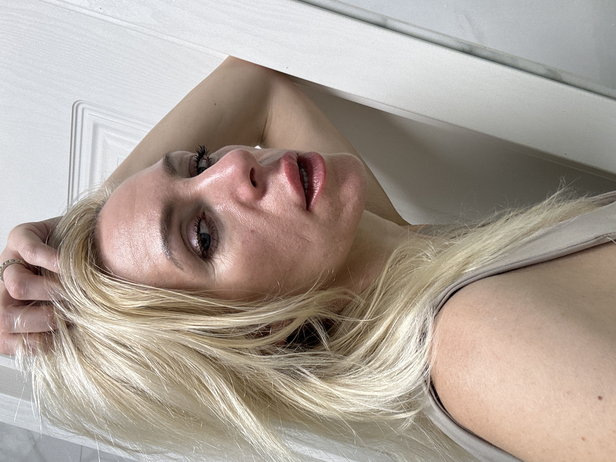 A close-up, medium shot captures a fair-skinned woman with blonde, tousled hair indoors. Her head is tilted back and to the right, with her right arm bent and resting behind her head, obscuring part of her face and hair. She is wearing a sleeveless, light-colored top. The background consists of a white wall with a door frame, casting subtle shadows that suggest soft lighting. A ring is visible on her right index finger. The woman's facial features are partially visible, with her mouth open as if speaking or about to speak. Her skin exhibits texture and some visible pores, with a light application of makeup evident on her lips and eyes. The scene is devoid of any specific landmarks or context that would indicate a location in any city.