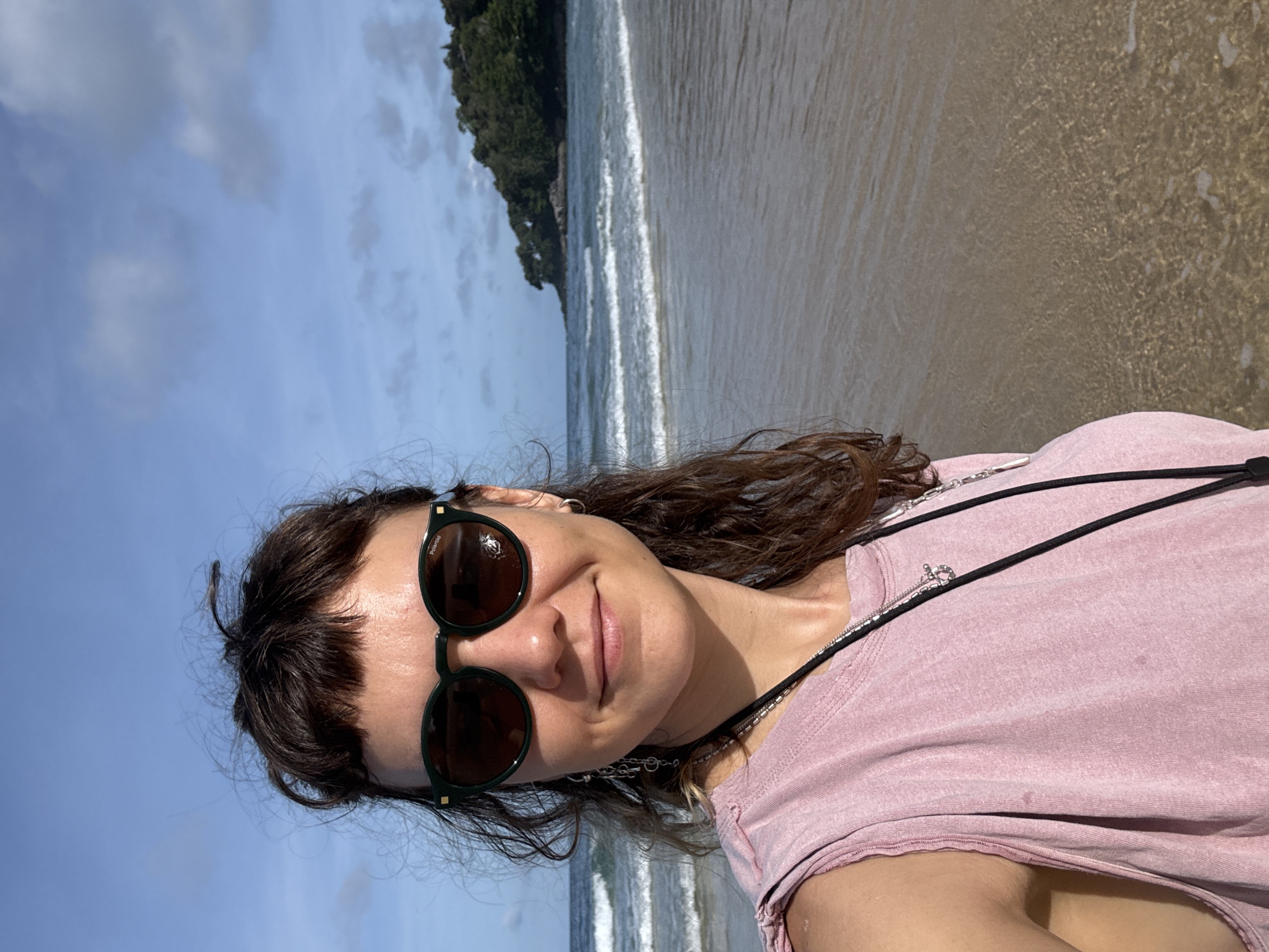 A young woman with dark, curly hair and bangs is taking a selfie on a beach. She is wearing sunglasses with dark lenses and a light pink, sleeveless shirt. She has on a necklace with a silver pendant. The ocean is behind her with gentle waves rolling onto the sandy shore. In the distance, a lush, green, tree-covered headland is visible. The sky is blue with scattered white clouds. The overall scene suggests a sunny day in a tropical coastal environment, likely Karon Beach, Thailand.