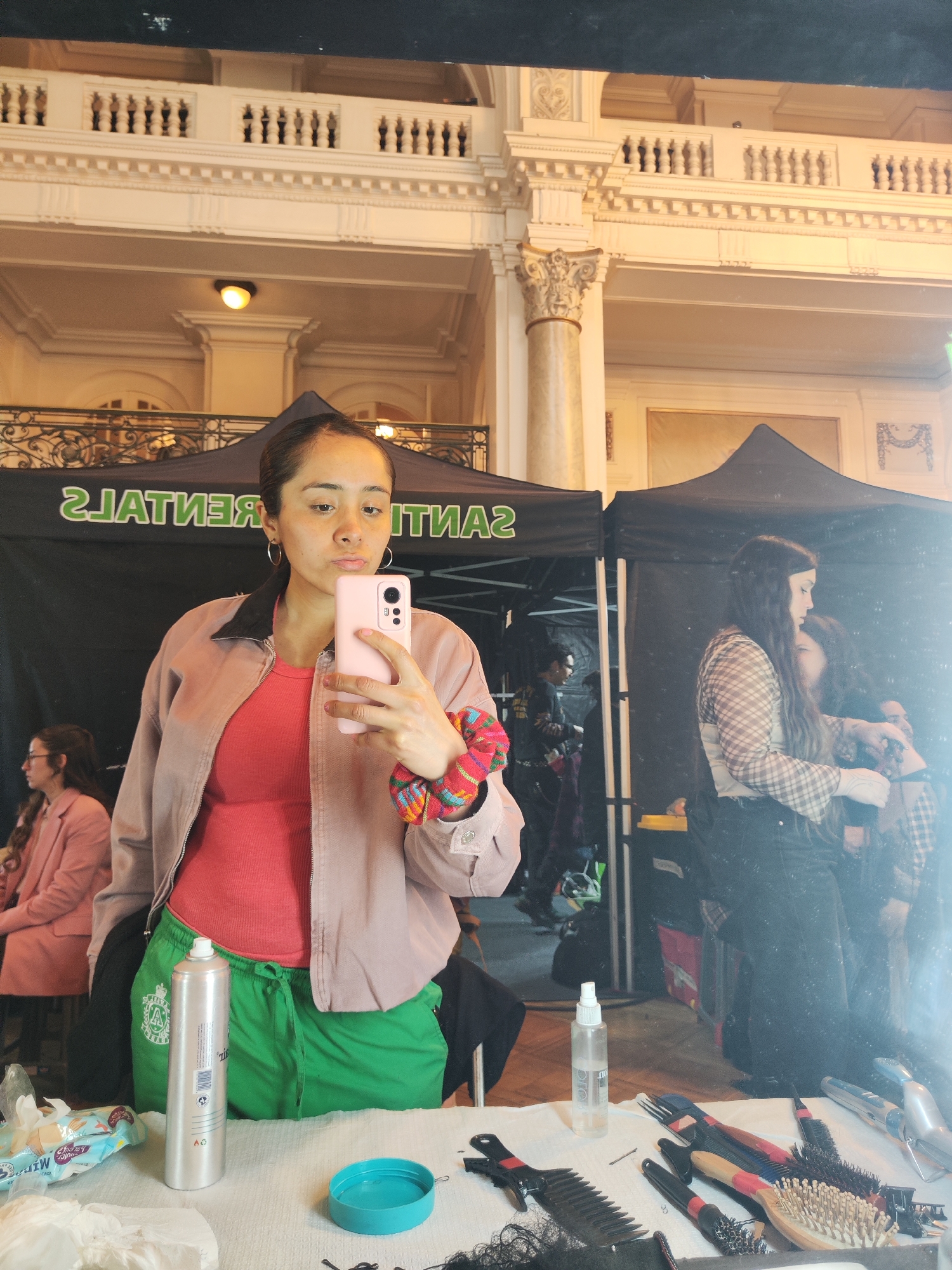 A medium shot captures a young woman in a pink jacket and coral top taking a selfie in a mirror. She has dark hair pulled back and is wearing hoop earrings. Her left arm is bent, holding a pink phone. A colorful scrunchie is on her right wrist. She is wearing bright green pants. In the background, a tent with the inverted text "RENTALS" is visible, suggesting a professional or event setting. To the right, a woman with long dark hair in a plaid shirt and jeans is reflected in the mirror, her hands near another person's hair, indicating a hair styling or makeup station. Other individuals and equipment are faintly visible in the background. The architecture in the upper portion of the image, with ornate columns and balconies, suggests a grand building, possibly a historic venue within Santiago, Chile. The overall scene appears to be a backstage or preparation area for an event.