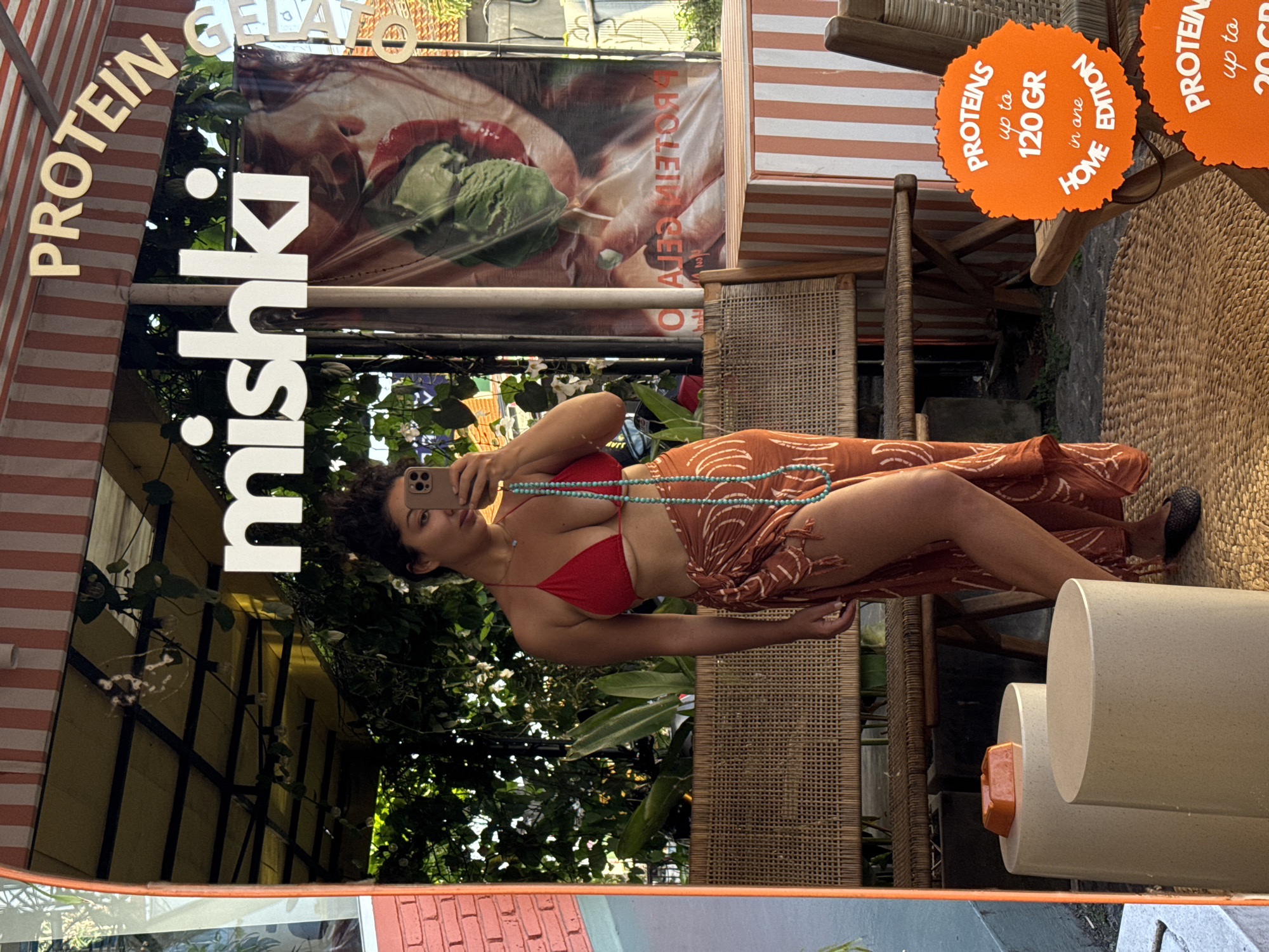 Here is a forensic description of the image:
The image is a reflection in a store window. The sign "mishki," with "PROTEIN GELATO" at the top, is visible. A woman in a red bikini top and orange sarong is visible in the reflection. She is holding up a phone and taking a picture of herself. Behind her is outdoor seating. Bright orange signs advertising "PROTEINS up to 120 GR in one HOME EDITION" and "PROTEIN up to 20 GR" are attached to some seating. The background has trees and foliage. A banner of gelato is visible on top of the reflection of the woman. The window is situated within a structure with alternating red and white stripes.
This location is likely in North Kuta, Indonesia.