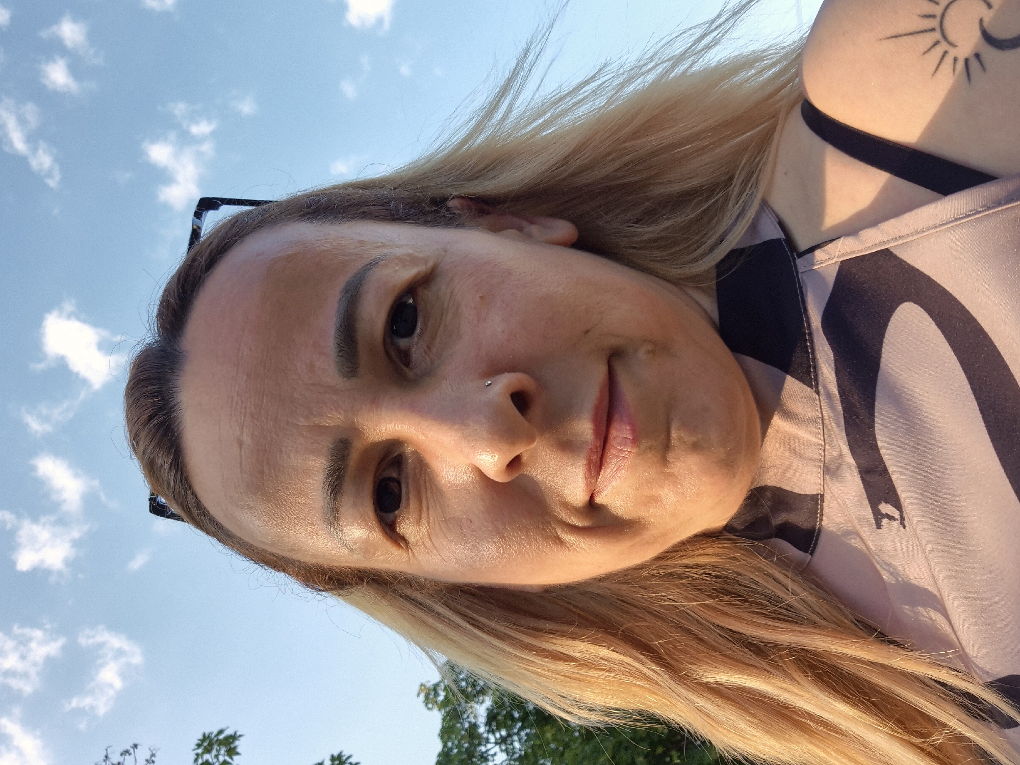 A medium close-up, upward-angled self-portrait depicts a light-skinned individual with long, blonde hair, smiling slightly and looking directly into the camera in the city of Unknown, Unknown. Dark-rimmed eyeglasses are perched on top of their head, partially obscured by hair. The forehead shows horizontal lines. Their dark brown eyes are open, and the nose features a small silver stud piercing the septum. The lips are lightly colored and slightly parted in a gentle expression. The person's left side (camera's right) of the face and hair is brightly illuminated by warm, directional sunlight, creating highlights and casting some areas of the right side (camera's left) into soft shadow. They are wearing a collared top or shirt with a light, possibly beige or pale pink fabric, adorned with a dark, abstract, wave-like pattern. A distinct stylized letter 'D' is visible on the patterned fabric below the chin. A dark strap from an undergarment or tank top is visible over the left shoulder (camera's left). On the upper left arm/shoulder, a black outline tattoo of a sun with rays and a crescent moon beneath it is visible. The background consists of a clear blue sky with scattered white cumulus clouds, and dark green foliage from trees is visible in the upper-left corner of the frame.