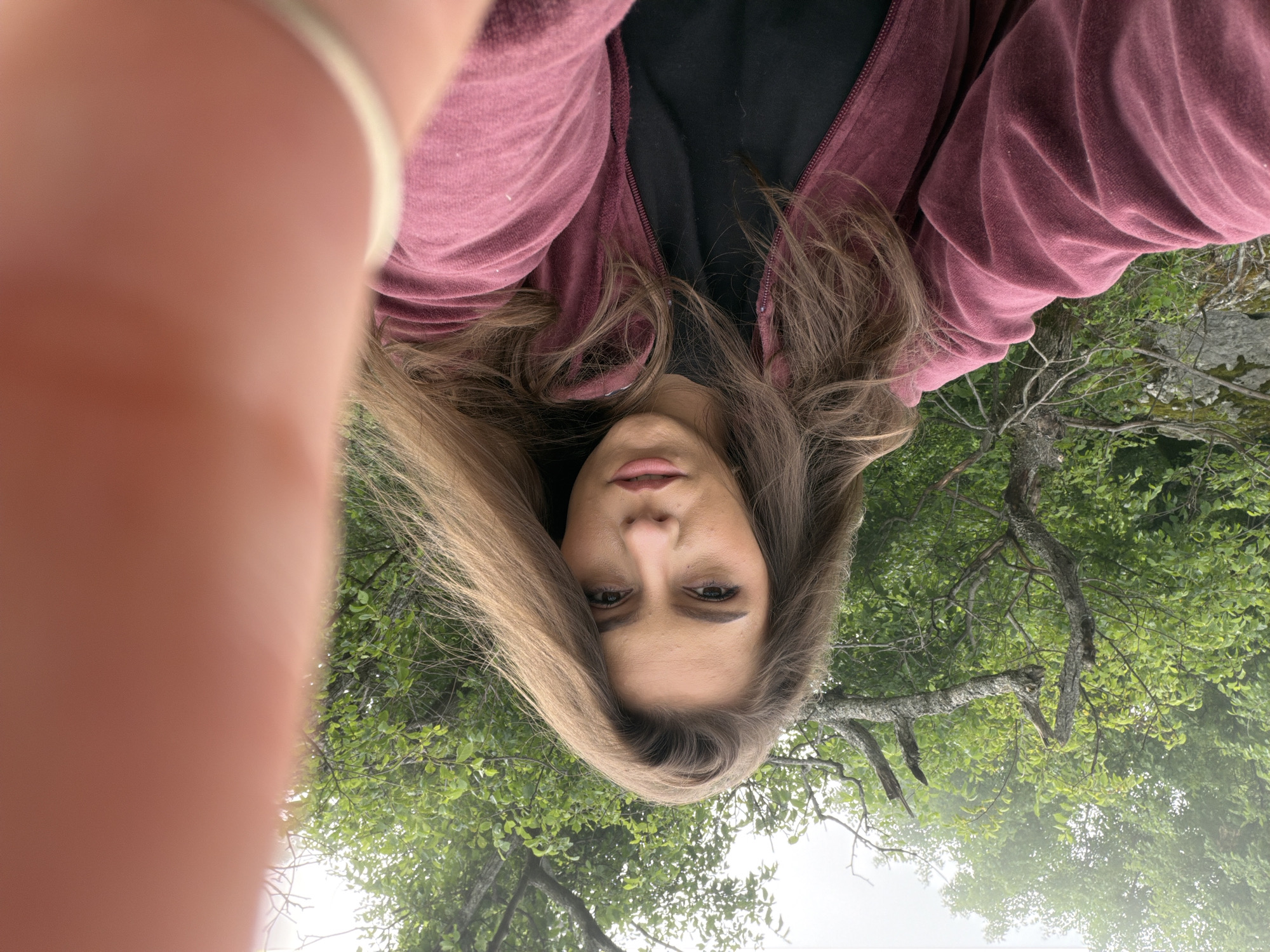 The image displays a woman in an unusual, inverted orientation, looking directly at the viewer. Her face is centered, framed by long, light brown hair that appears to fall upwards in the current frame. Her lips are slightly parted, and she has a neutral expression. She is wearing a dark red or maroon zippered jacket over a black top. A blurred finger or thumb partially obscures the lower-left portion of the image, suggesting a selfie taken at close range.
The background is filled with lush green foliage, featuring numerous tree leaves and branches, indicating an outdoor, natural setting such as a forest or park. The perspective suggests the camera was pointing upwards, towards the trees, before the image was rotated. The lighting is soft and diffused, characteristic of an overcast day or light filtering through a dense tree canopy. No text is visible in the image.
