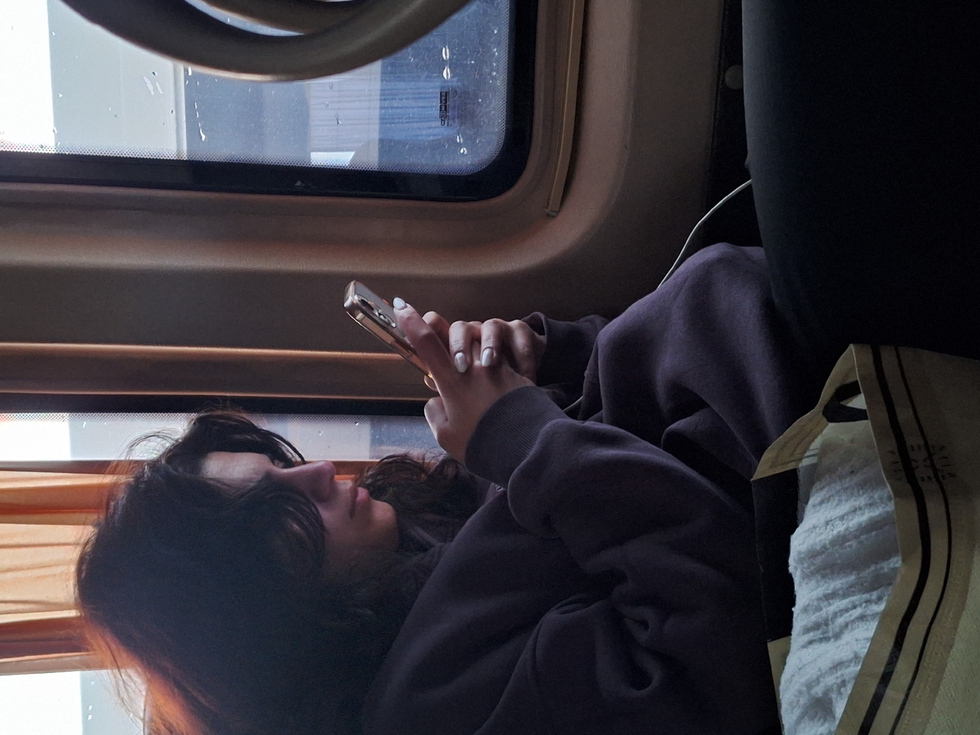A young woman is seated on a train, looking at her phone. She has long, dark curly hair and is wearing a dark purple sweatshirt. Her hands are holding a gold-colored smartphone. To her right is a white bag with dark stripes. Behind her is a black headrest. The train is traveling past windows with a view of buildings. The image provides no information about the city of Anipemza, Armenia.