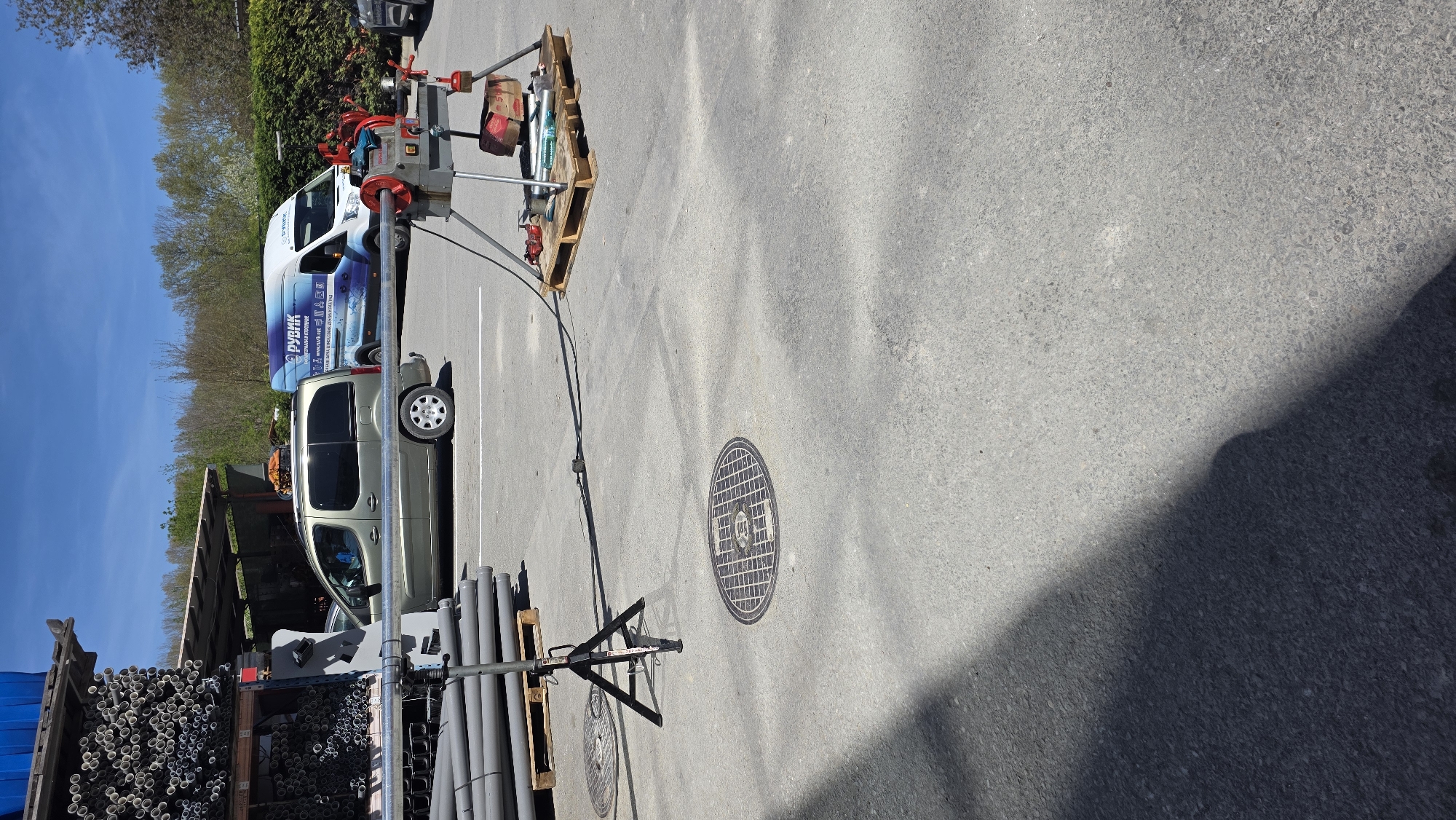 The image shows an outdoor industrial scene in Varna, Bulgaria. It appears to be a storage or work area, possibly related to plumbing or construction, evidenced by the presence of pipes and equipment.
The scene features:
- Two vans parked in marked spaces; one is white with blue graphics and the text "РУБИК" visible, while the other is a silver/grey minivan.
- A large number of pipes are stored on a rack or shelf, with both small and large diameter pipes visible.
- Equipment, including a threading machine mounted on a tripod, is situated nearby. A pallet with other tools is also present.
- A manhole cover is visible on the pavement.
- The ground is paved with asphalt.
- The background includes trees and shrubs under a bright blue sky.