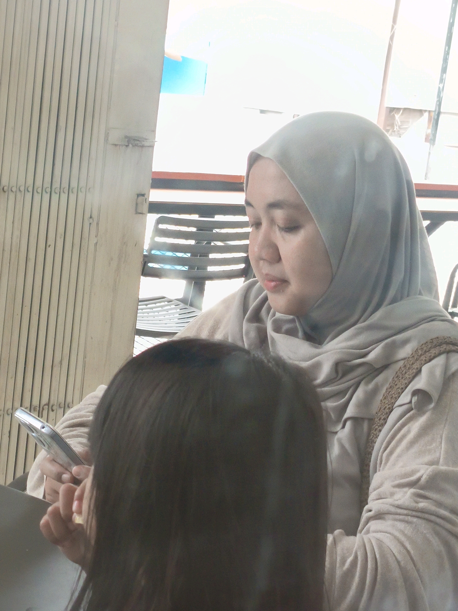 In an outdoor setting, a woman wearing a light gray hijab and a beige sweater is seated, looking down at her smartphone. Her left hand is holding the phone, while her right hand is visible in front of her. The background features a closed, light-colored metal accordion door on the left and blurred chairs and a bright sky on the right. A child's dark hair is in the foreground, obscuring part of the woman and her phone. The scene appears to be in Indonesia.