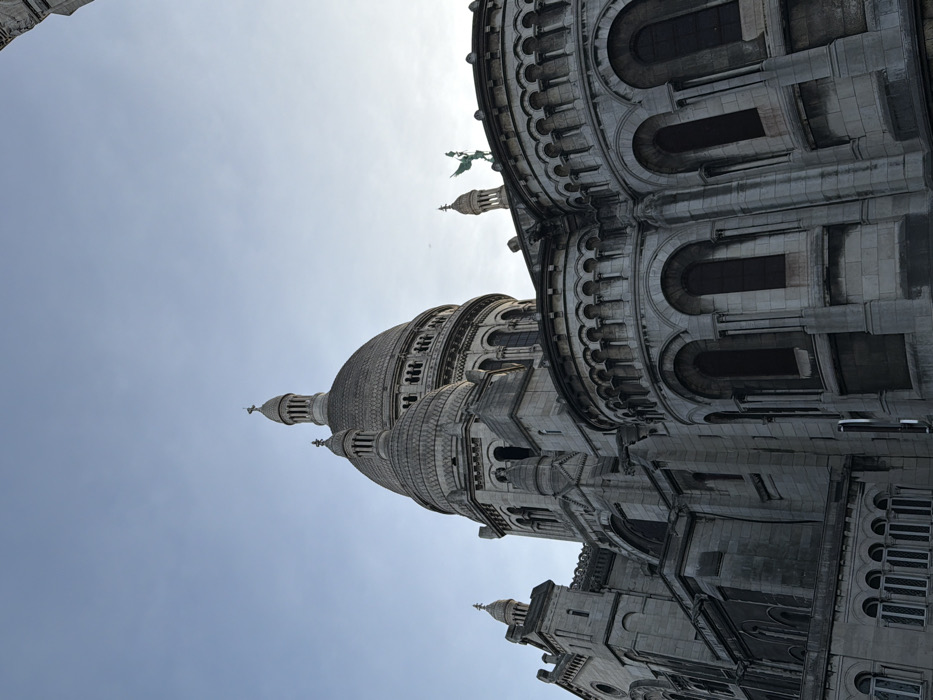 The image presents a low-angle perspective of the Sacré-Cœur Basilica, located in the Montmartre district of Paris, France. The monumental structure, constructed of light-colored stone, occupies the majority of the frame against a clear, pale blue sky. A prominent central dome, capped by a lantern and a cross, rises in the upper center, accompanied by smaller domes and cupolas on either side. The architecture exhibits numerous arched windows and decorative archways, supported by columns and pilasters. Intricate stonework, including carved details and corbel tables, is visible across the building's facade. On the right side of the roofline, a green-patinated statue depicting a winged figure stands atop a structural element. No individuals, actions, or interactions are present in the scene.
