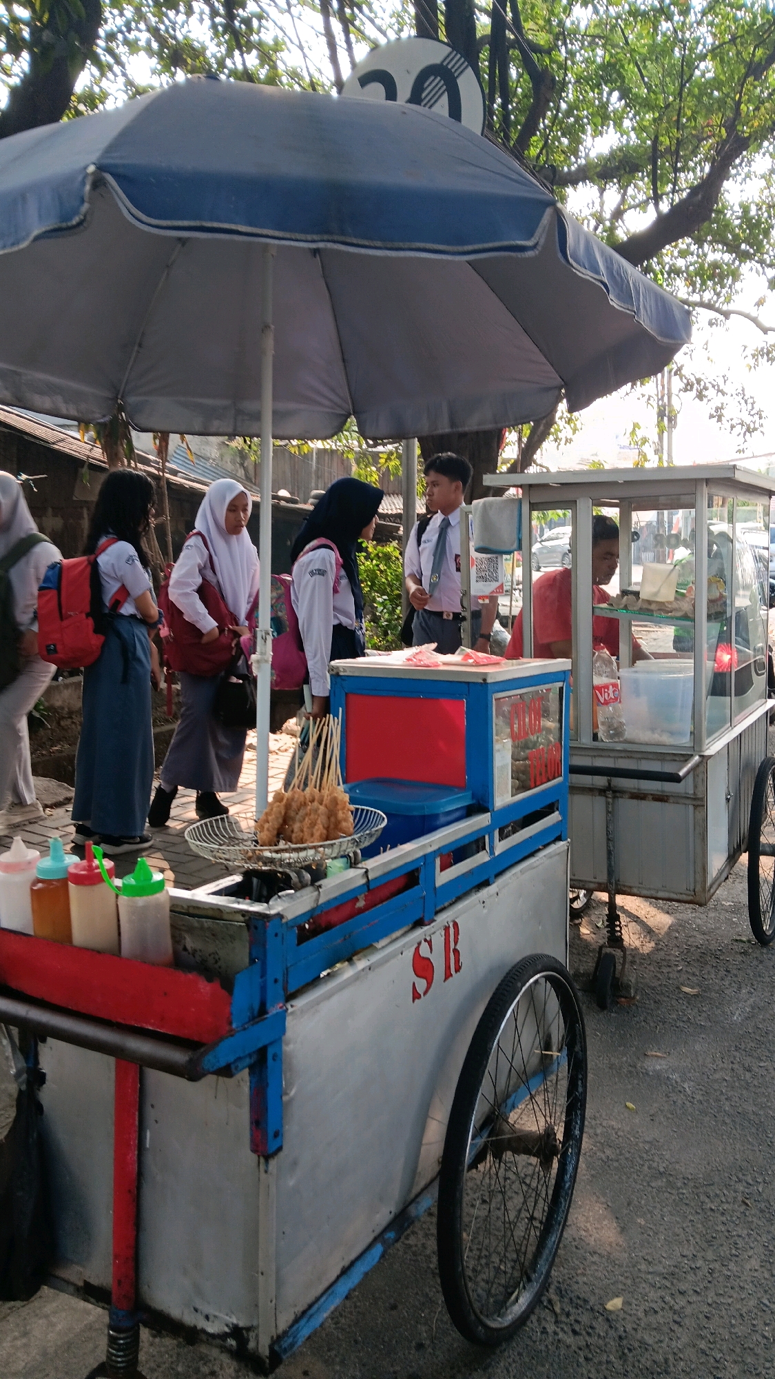 In South Jakarta, Indonesia, on a sunlit street, a food cart vendor serves customers under a large blue and gray umbrella. The cart is divided into two sections: the front section displays skewers of fried food in a white basket, along with three condiment bottles, and a red and blue cabinet. The rear section is a glass display case with a vendor inside, wearing a red shirt and serving food to customers.
Several individuals, appearing to be students in school uniforms, are gathered around the cart, some interacting with the vendor. One student wears a white shirt, gray skirt, and red backpack. Another wears a white shirt, black hijab, and pink backpack. A male student in a white shirt, tie, and gray pants stands near the rear of the cart. The surrounding environment suggests a typical urban street scene, with a road surface, trees providing shade, and other vehicles visible in the background. A speed limit sign is visible in the upper portion of the image.