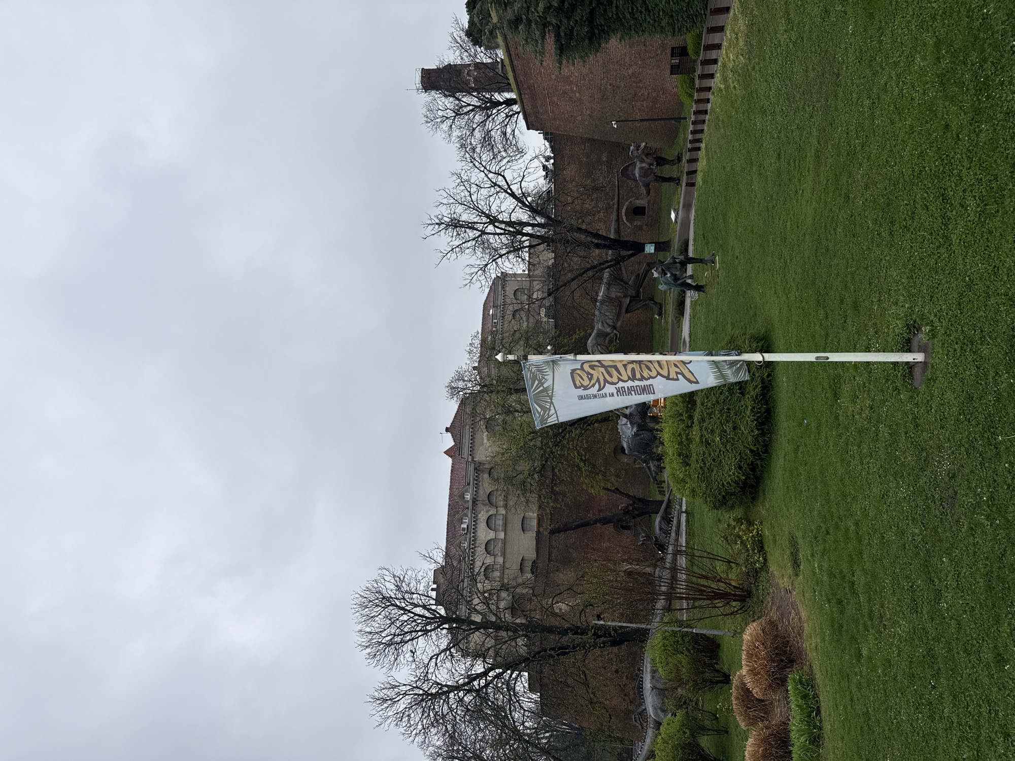 The image presents an outdoor scene at Kalemegdan Park, Belgrade, Serbia, under an overcast sky. A lush green lawn with scattered clover and patches of dried ornamental grasses occupies the foreground. A white flagpole, supporting a light-colored banner with inverted text and graphics, stands centrally. Several large, grey-colored dinosaur models populate the midground, including a bipedal carnivore, a spined bipedal dinosaur, and other partial figures, positioned on the grass and near a paved path. A prominent, tall reddish-brown brick fortress wall, characteristic of Kalemegdan, extends horizontally across the scene, with numerous bare-branched trees scattered in front of and behind it. Above the fortress wall, a large, light-colored building with a red-tiled roof and arched windows is visible, along with a narrow, cylindrical brick tower on the far right. The sky is uniformly light grey and heavily clouded. No people are present in the frame.