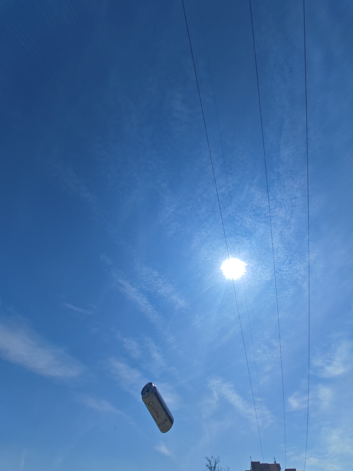 The image displays a bright, clear blue sky with scattered, wispy white clouds. The sun is visible in the upper right quadrant, emitting a strong glare. Four thin, dark parallel overhead wires traverse diagonally across the upper right portion of the frame. In the lower-middle left, a silver-colored "Monster Energy Zero Sugar" can is observed in mid-air, oriented diagonally upwards and to the right. A partial view of a building's roofline featuring multiple dark structures and a bare tree branch is visible at the extreme bottom right edge of the frame. The scene is located within the city of Shumen, Bulgaria, depicting an urban or suburban skyward view. No people are visible.