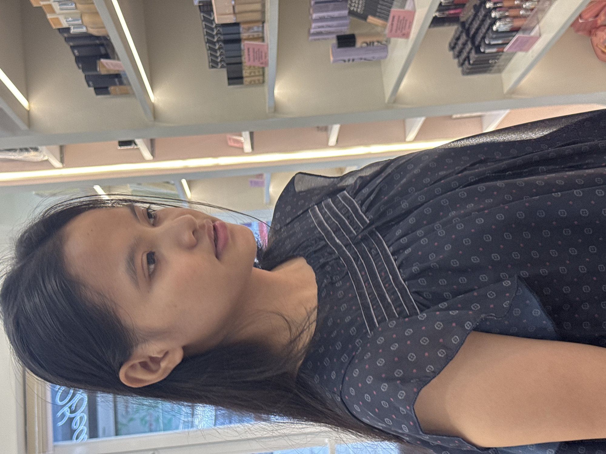 A young Asian woman with long, dark hair is standing in front of a shelf stocked with cosmetic products. The products are arranged neatly on multiple shelves, suggesting a retail environment, likely a makeup store. The woman is wearing a dark, patterned dress with a detailed bodice. Through the window behind her, a sign with the word "OPEN" is visible, hinting at the store's operational status. There is no specific indicator that the image was taken in Davao City, Philippines.