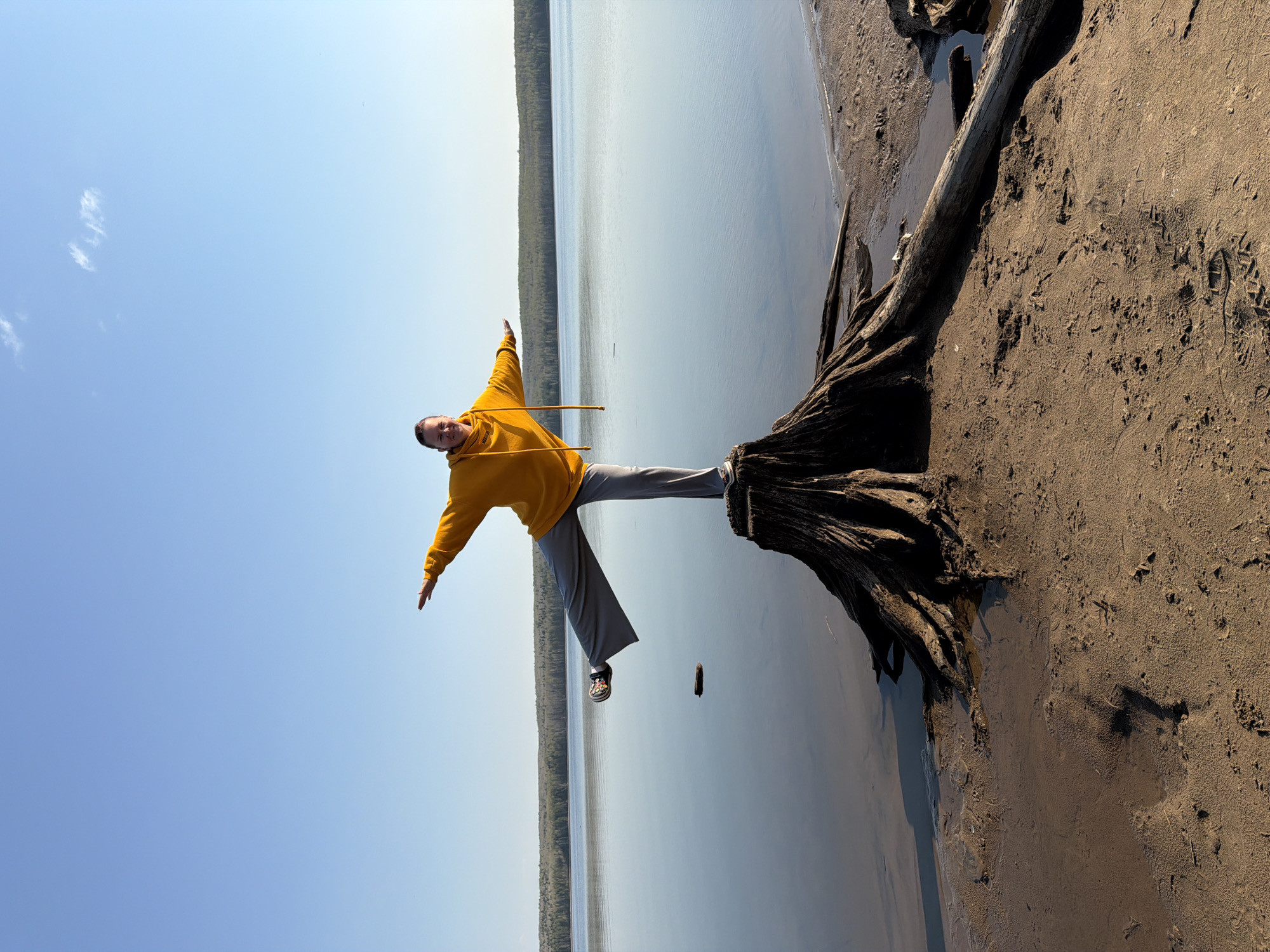 A person stands on a tree stump near a body of water, likely in Chaikovsky, Russia. The person wears a yellow hoodie and gray pants, extending their arms outward. The stump sits on a sandy bank adjacent to the water. Beyond the water, a treeline is visible under a blue sky.