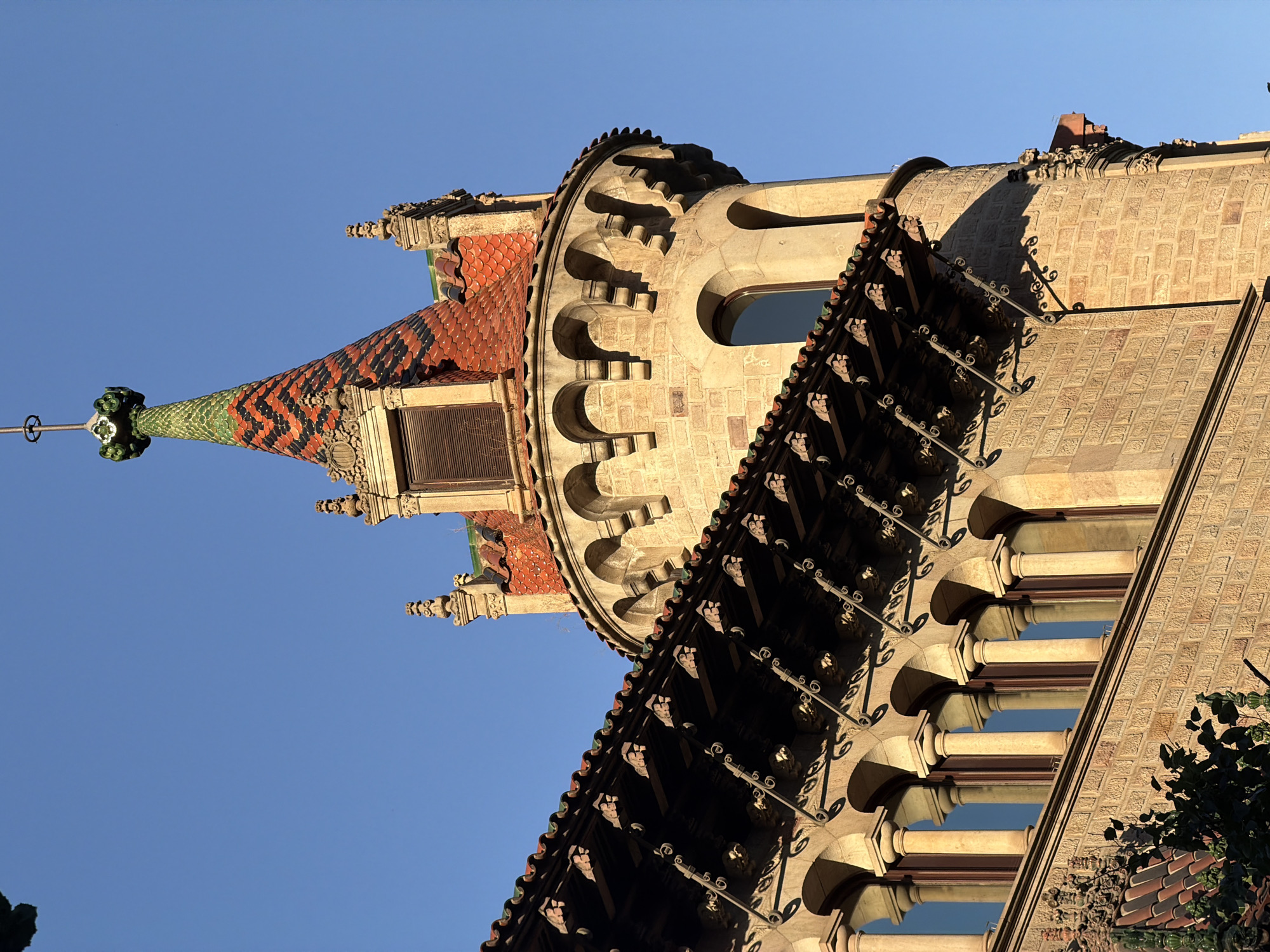 The image shows a close-up view of a turret on a building, set against a clear blue sky. The turret has a conical roof covered in red and black patterned tiles, with green accents near the top. Below the roof, the turret is constructed of light-colored stone blocks and features arched windows and decorative stonework. To the left of the turret, a section of the building's roof is visible, supported by a series of arches and columns, with intricate metalwork and carved details. The building's facade is made of brickwork, with some decorative elements. The lighting suggests it is either morning or late afternoon, casting shadows across the architecture. The building appears to be a historical or architecturally significant structure in Barcelona, Spain. The specific location is Casa Vicens, one of Antoni Gaudí's early works, located in the Gràcia district of Barcelona. There are no people or actions visible in the image.