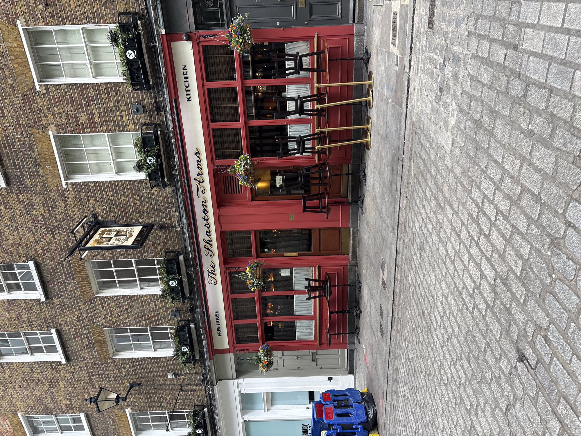 The image depicts the facade of "The Shaston Arms" pub in London, United Kingdom. The pub is painted red with a white awning that reads "The Shaston Arms" and "KITCHEN FREE HOUSE" in black lettering. It has multiple window sections and a door. Three tables with chairs stand outside the pub on the cobblestone street. The pub is located next to a brick building with multiple windows, each adorned with flower boxes. A street lamp is visible on the brick building. A blue barrier sits adjacent to the building on the street.