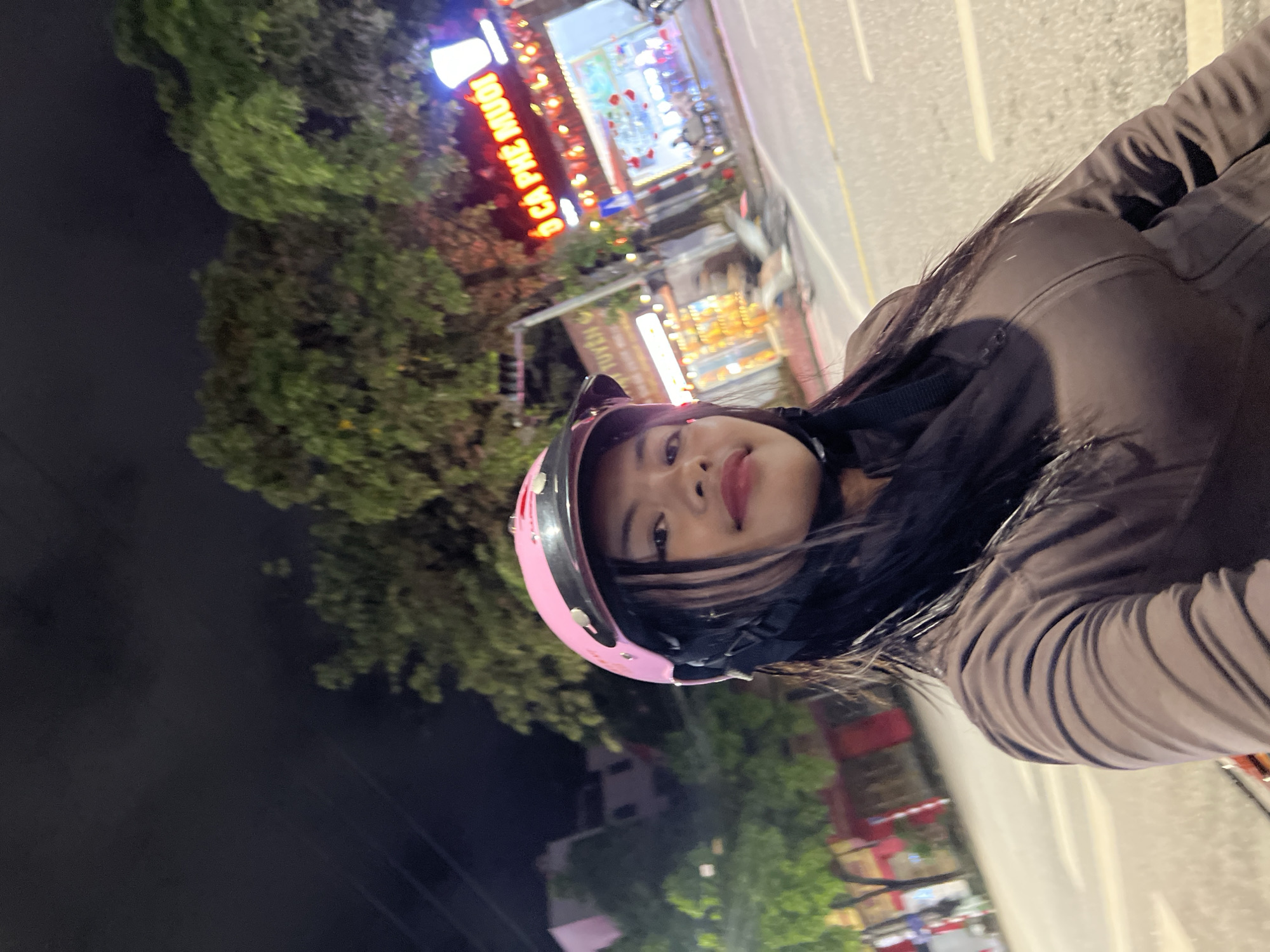 A young woman wearing a pink helmet and a brown jacket is taking a selfie at night in Minh Xuân Ward, Vietnam. The background shows a street with trees, buildings, and neon signs, indicating an urban setting. One sign reads "CÀ PHÊ MUỐI" in large, illuminated letters, suggesting a coffee shop. The road appears wet, possibly from rain. The overall scene depicts a casual moment captured during nighttime in a Vietnamese city.