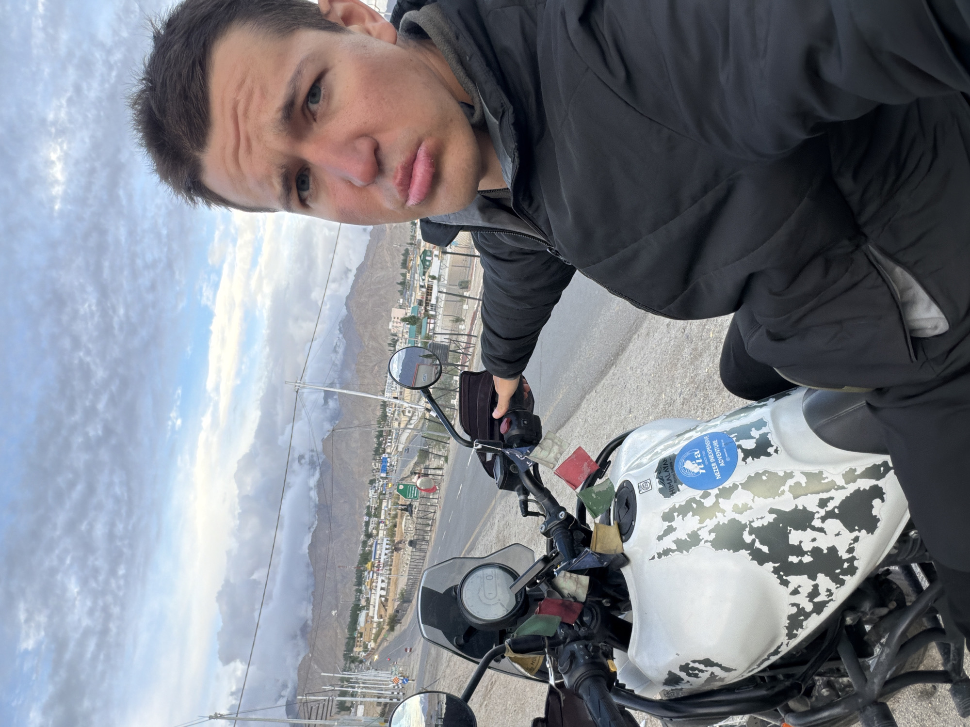 A young Caucasian man, wearing a black jacket, is seated on a white motorcycle with camouflage accents and a blue sticker on the fuel tank. He is looking at the camera with his mouth slightly pursed. The motorcycle is parked on the side of a road, with a town and mountains in the background. The sky is cloudy. The location is Spituk, India.