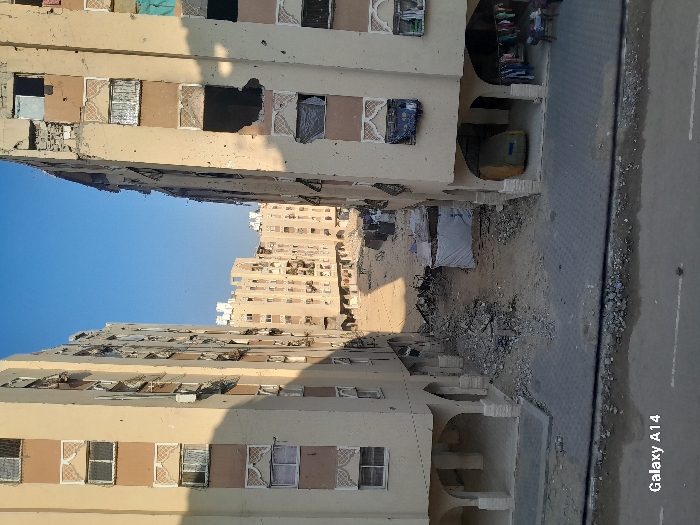 The image depicts an urban landscape in Khan Yunis, Palestinian Territory, characterized by residential buildings showing significant damage and debris. Two multi-story apartment blocks, primarily beige with brown accents and patterned window frames, frame a central area. The building on the right exhibits substantial damage, including large holes through multiple floors, shattered windows, and external pitting, with several window openings covered by tarps or cloths of various colors (blue, light blue, dark grey). The building on the left appears less severely damaged, though multiple windows are open or have external air conditioning units.
Between these two foreground buildings, a wide unpaved area is filled with extensive rubble, fragmented concrete, and disturbed earth, suggesting demolition or severe structural collapse. Towards the right side of this central area, a small, makeshift structure or tent, covered in white tarpaulin, is present. In the background, additional multi-story residential buildings are visible, also exhibiting signs of wear and possible damage.
At the ground level, a paved sidewalk constructed from interlocking grey pavers runs across the bottom of the frame, separating the buildings from a debris-strewn asphalt street. Under an archway on the ground floor of the right building, a yellow vehicle is parked, covered by a tarp. Adjacent to it, laundry is hung on a line, and two individuals are seated on low chairs, suggesting a functional living or resting area despite the surrounding destruction. The scene is illuminated by clear daylight under a bright blue sky. A "Galaxy A14" watermark is visible in the lower left corner.