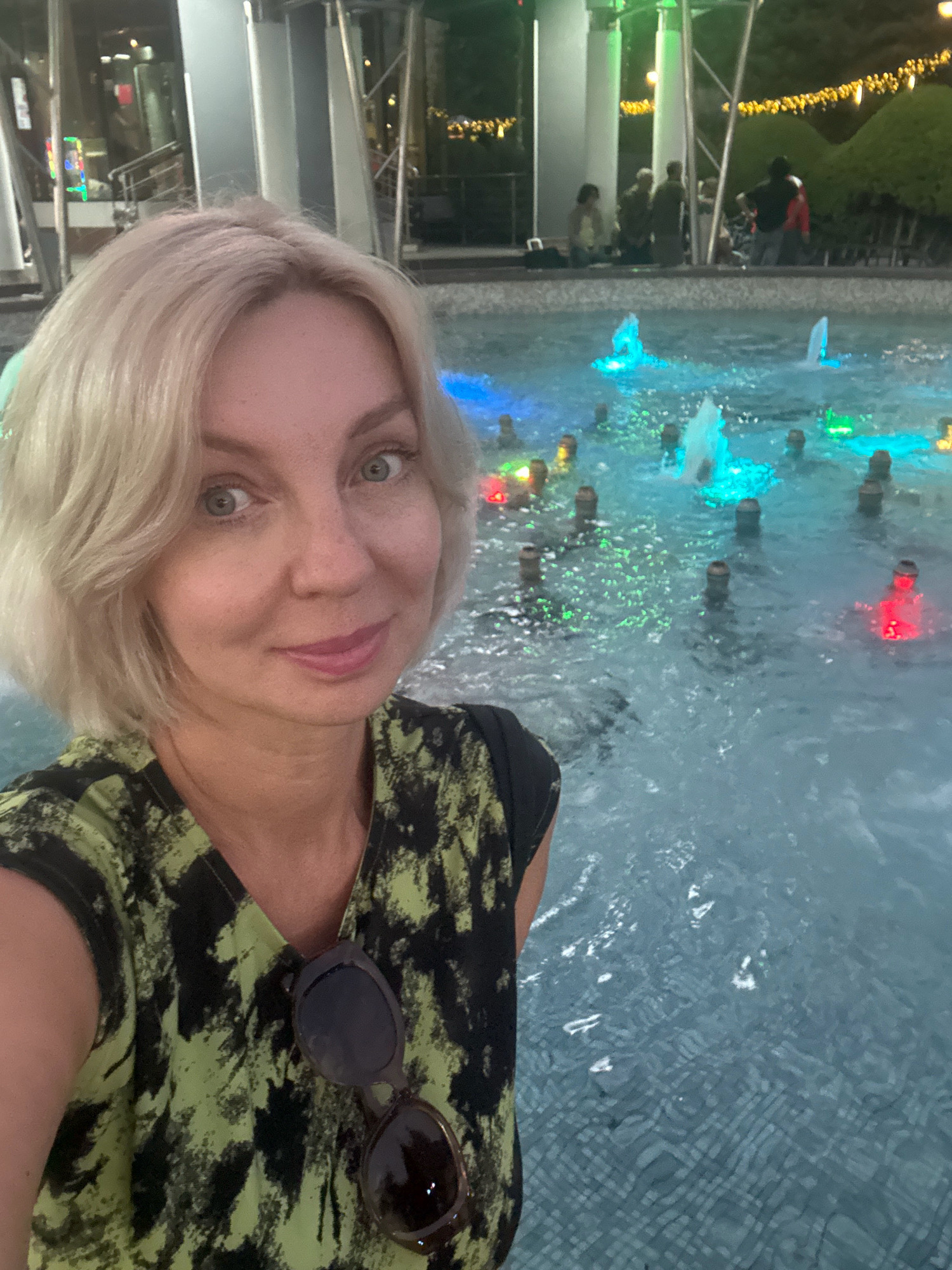 A selfie of a woman with short blonde hair and green eyes is taken in front of a fountain with illuminated water jets. The woman is wearing a green and black patterned t-shirt and sunglasses are hanging from her neckline. In the background, several people are gathered around the fountain, and decorative lights are visible above them. The fountain appears to be located in a public outdoor space, possibly a park or promenade, within the city of Sochi, Russia.