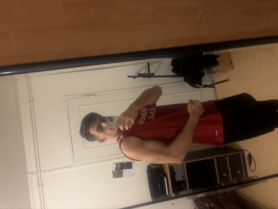 A man in a maroon sleeveless shirt and black pants poses in front of a mirror, flexing his bicep while holding a smartphone to take the photo. He stands inside a room with white walls. A white door is behind him. Above the door hangs a chin-up bar with workout gear. Below, a small cabinet sits beside a small desk. The mirror has a dark frame.
