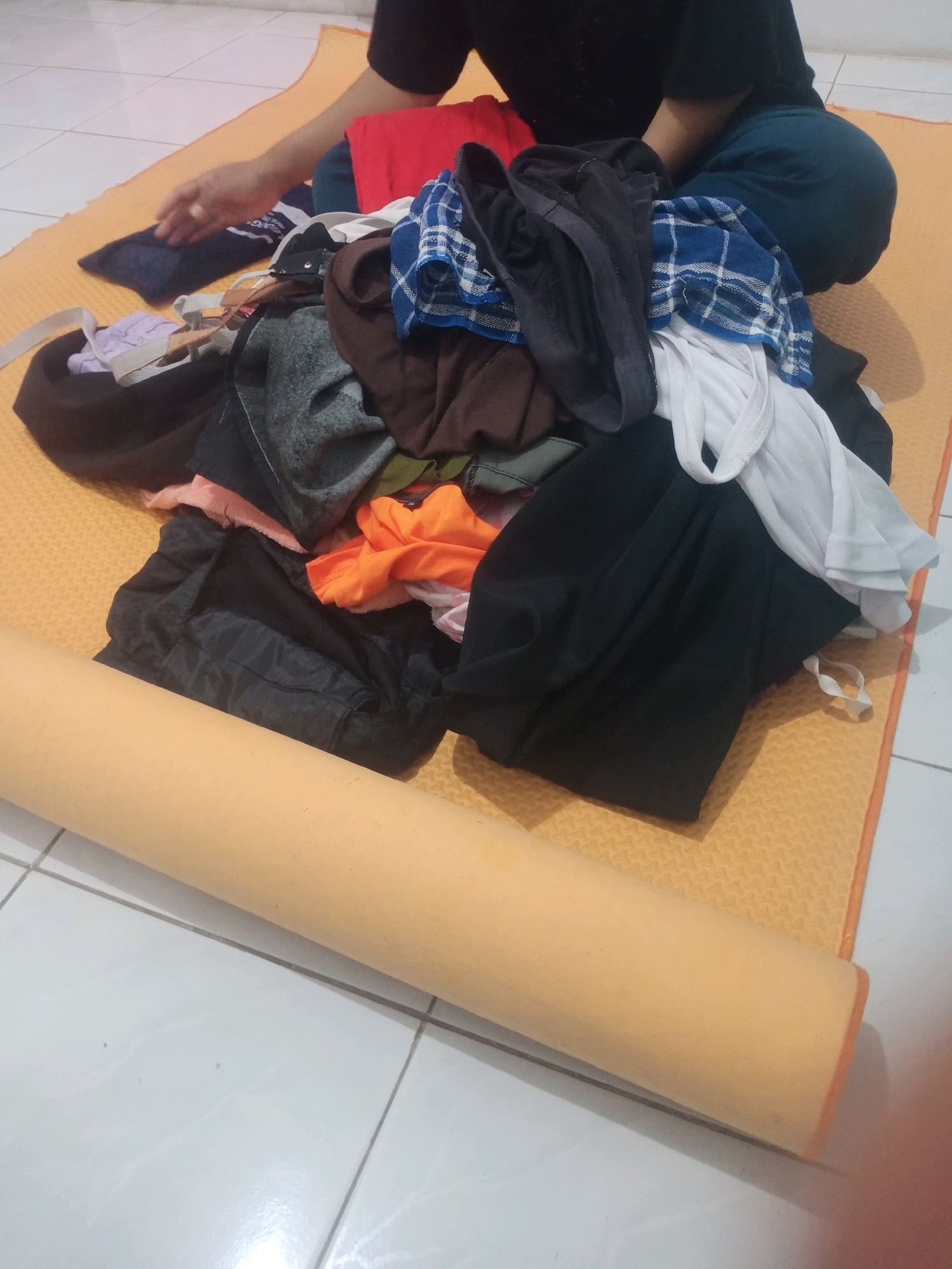 The image depicts a person, likely male based on clothing, sitting on an orange yoga mat on a tiled floor. The person is wearing a black t-shirt and blue pants. The person's hands are visible, interacting with a pile of folded clothes on the mat. The clothes are varied in color and fabric, including a blue and white checkered shirt, a red shirt, a black garment, and a white tank top. A rolled-up beige yoga mat is positioned in the foreground, partially obscuring the lower portion of the scene. The lighting suggests an indoor environment, possibly a bedroom or living area. The presence of folded laundry and a yoga mat suggests a domestic setting. The location of Bandar Lampung, Indonesia, is not directly indicated by any visual cues within the image.