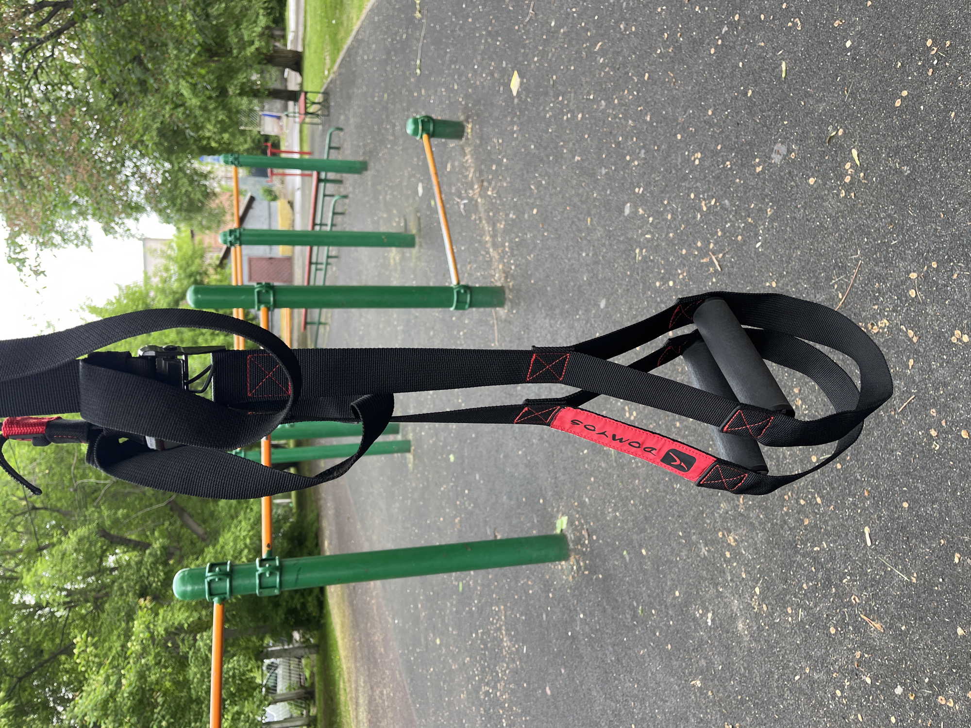 A close-up shot focuses on a black suspension trainer with red trim, hanging outdoors on a cloudy day at a public exercise park. The trainer, featuring foam handles and adjustable straps, is the primary subject. In the background, green metal posts with orange horizontal bars, typical of outdoor gym equipment, are visible. The ground is a dark, textured surface, possibly rubberized, with scattered leaves and debris. Trees with green foliage are present in the distance, along with a building and a fence. The scene suggests a common urban outdoor fitness area.