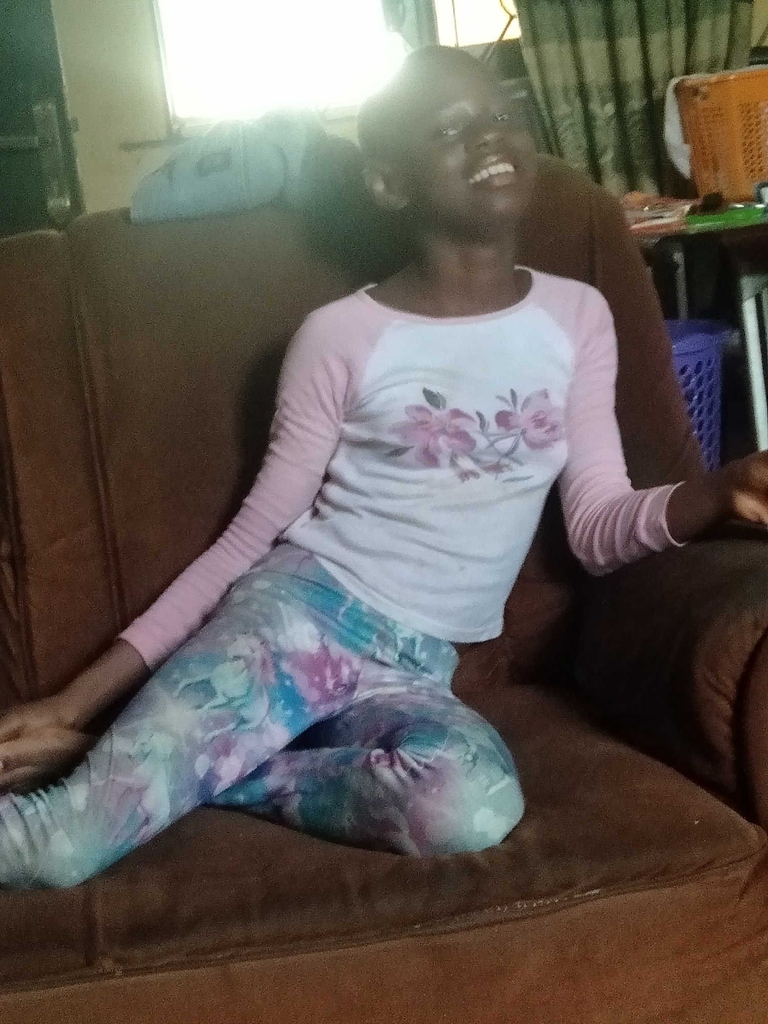 A candid, brightly lit medium shot captures a young, dark-skinned individual, likely a child or pre-teen, seated on a brown plush sofa in an indoor setting. The subject, positioned slightly off-center to the right, is looking upwards and laughing heartily with a wide, joyful smile, their head tilted back.
They are dressed in a casual outfit: a long-sleeved shirt with a white body and pink sleeves, featuring a delicate floral design of pink flowers and green leaves on the chest. Their lower body is covered by vibrant, tie-dye-patterned leggings in shades of blue, purple, and pink. Their arms are relaxed, resting on the sofa's armrests or by their sides.
The background includes a brightly illuminated area to the left, indicative of a window letting in natural daylight, which causes some overexposure. To the right, a dark green patterned curtain is visible, along with glimpses of household items such as a purple plastic basket. A light-colored cushion or pillow rests on the back of the sofa behind the subject's head. The scene suggests a moment of uninhibited joy during the daytime, likely in a living area in Shangisha, Nigeria. No visible text is present in the image.