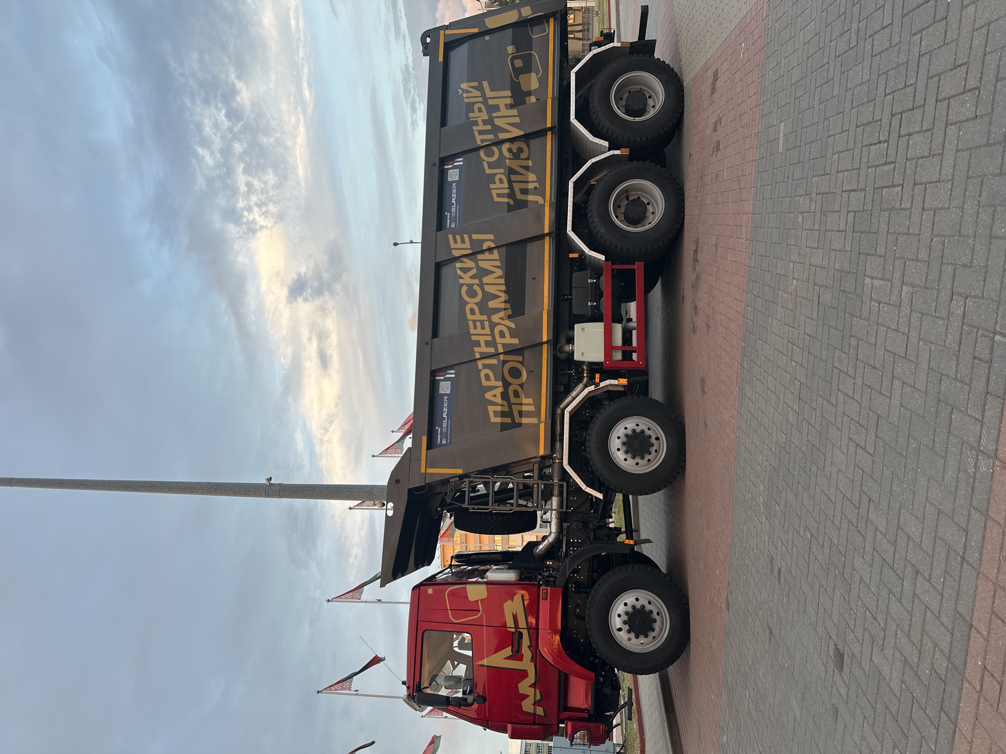 The image shows a side view of a large red and gray dump truck parked on a paved surface composed of interlocking bricks. The truck has the brand name "MAZ" on its side, and large text in gold on the gray dump body reads "ПАРТНЕРСКИЕ ПРОГРАММЫ" and "ЛЬГОТНЫЙ ЛИЗИНГ," which translates to "PARTNERSHIP PROGRAMS" and "PREFERENTIAL LEASING" in English. Several flags, appearing to be the national flag of Belarus, are visible in the background, suggesting the location may be in Minsk, Belarus, possibly near a government building or event space. The sky is cloudy with hints of sunset colors. There are no people visible in the image.
