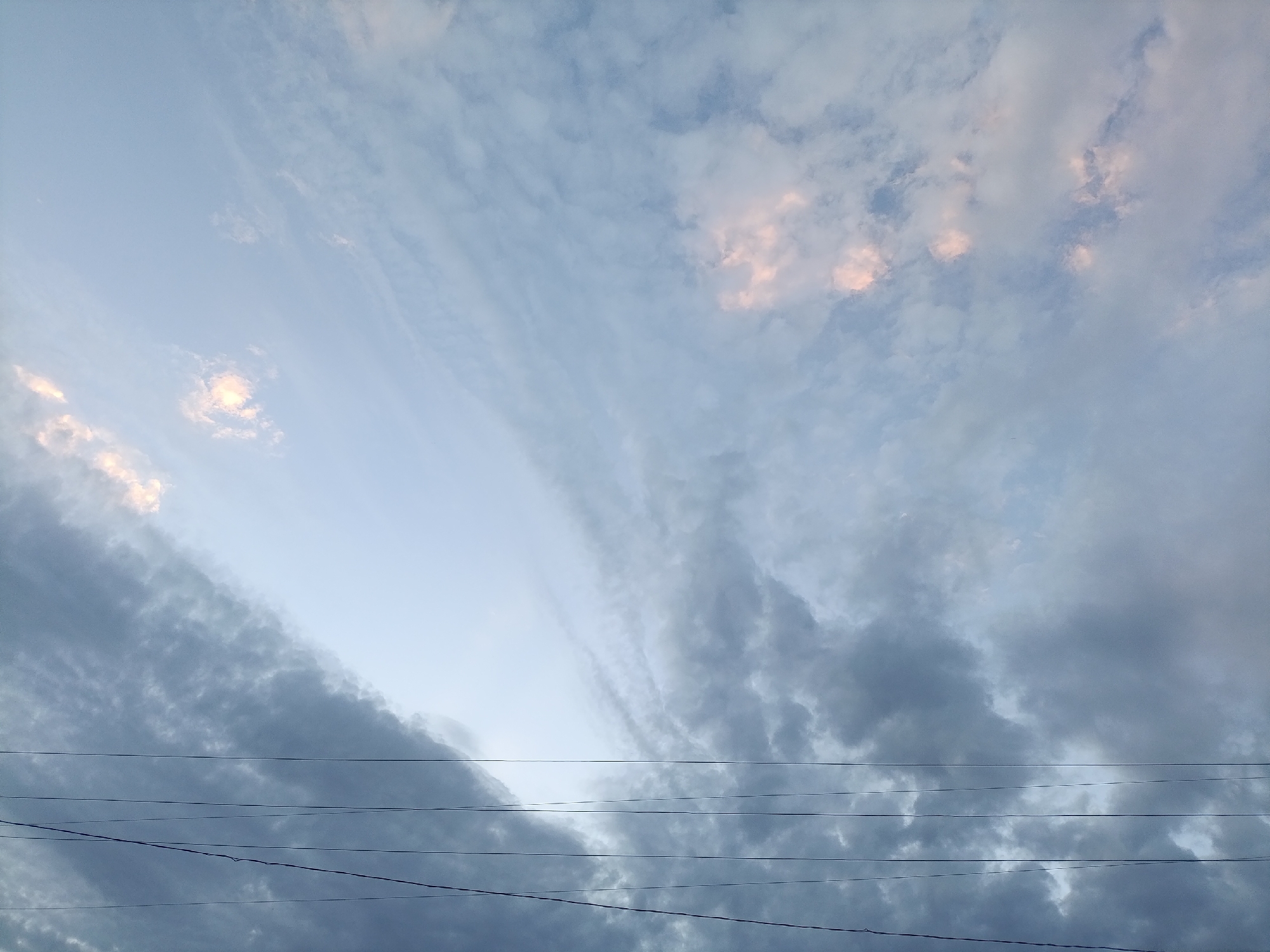 The image captures a wide-angle view of the sky above Kyzyl-Suu, Kyrgyzstan. The sky is a blend of soft blues and grays, with scattered clouds. Some of the clouds exhibit a subtle pinkish hue, suggesting the influence of dawn or dusk light. Multiple thin, dark wires stretch horizontally across the lower portion of the frame, indicating the presence of overhead power lines. These wires are distinct against the lighter sky and darker clouds, adding a man-made element to the natural scene. The overall impression is one of a serene, open sky with the subtle interruption of utility infrastructure.