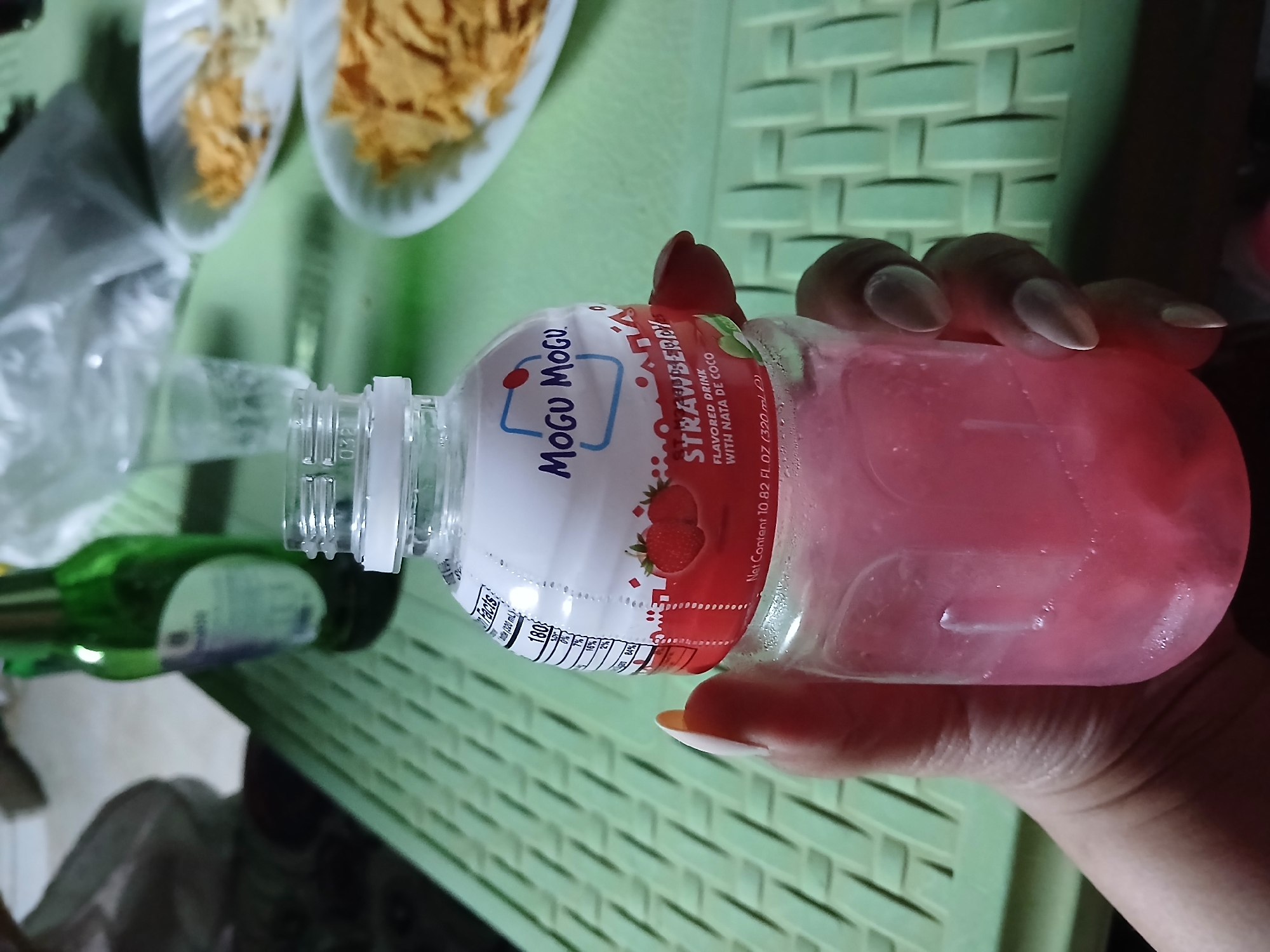 A close-up shot shows a hand holding a bottle of Mogu Mogu strawberry-flavored drink with nata de coco. The bottle is clear and filled with pink liquid. The label on the bottle features a drawing of strawberries and the Mogu Mogu logo. In the background, a green woven surface is visible, along with blurred images of a green bottle, a glass, and two white plates filled with chips. The scene is likely indoors, possibly in a casual setting. The location of Barza Municipality, Syria is not discernible from the image.