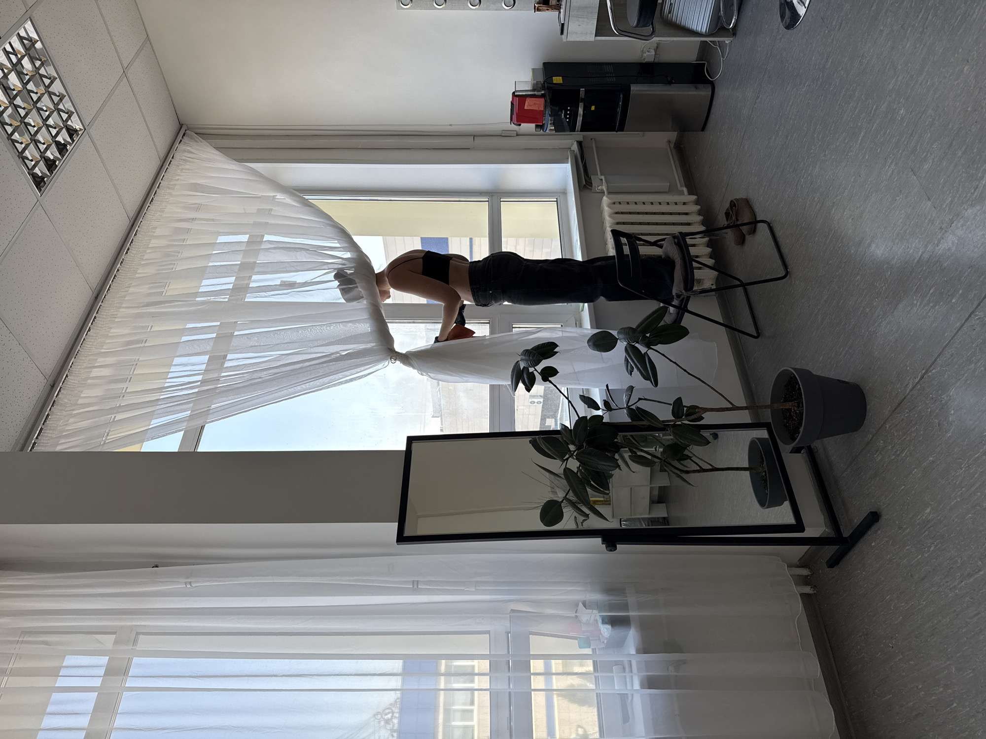 The image captures a scene inside a room in Izhevsk, Russia. A person stands precariously on a chair by the window, partially obscured by a white curtain. The person is wearing a black top and dark jeans, and appears to be adjusting or looking out from behind the curtain. Adjacent to the window, there's a tall, narrow mirror and a potted plant. In the background, office equipment and chairs are visible, with the room being lit by overhead ceiling lights. The flooring is a uniform grey, possibly linoleum or a similar material. A lower window and buildings outside are visible below the mirror.