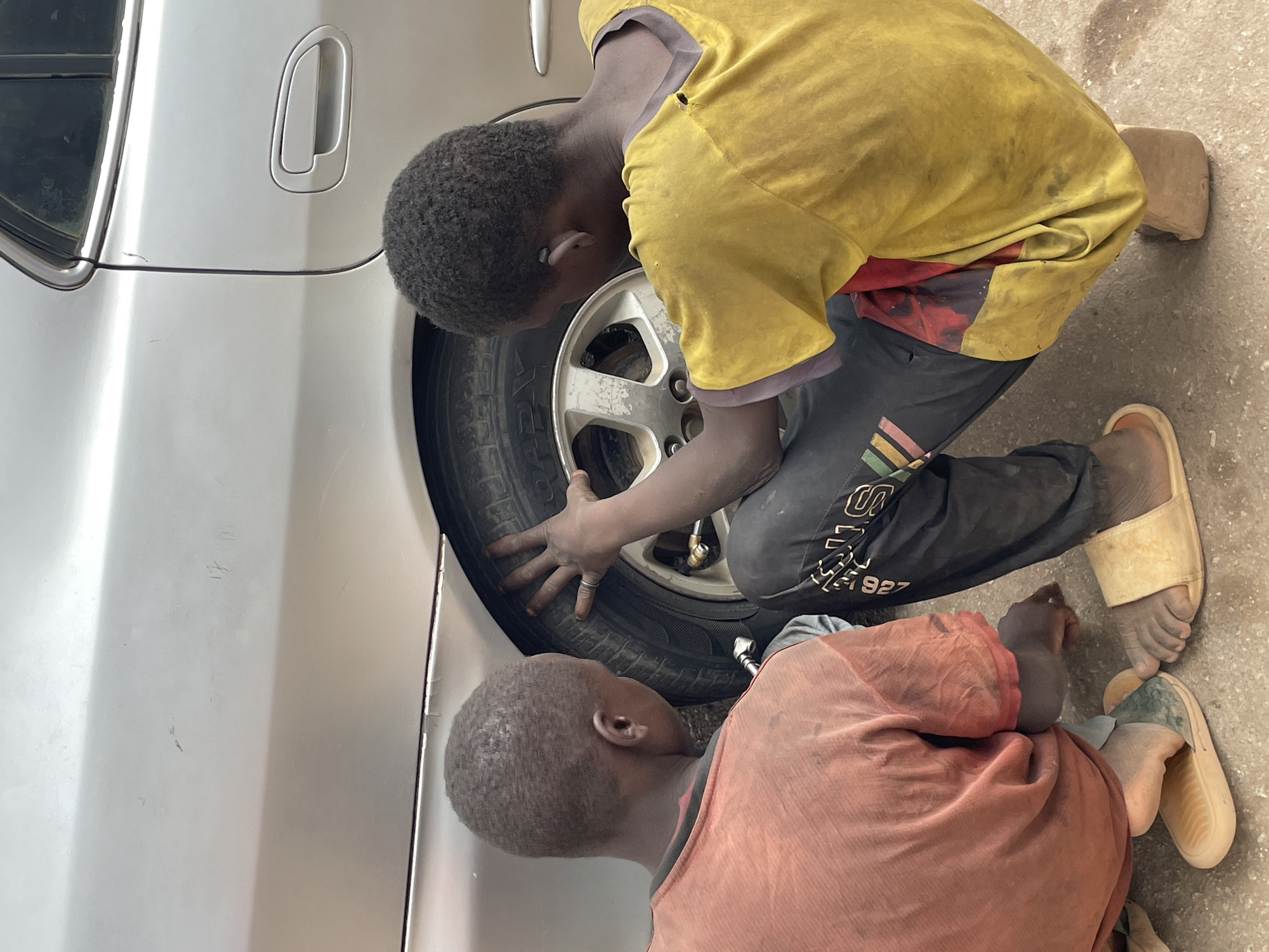 Two young boys are seen attending to a car tire in an outdoor setting, likely a repair shop or roadside. The image is taken from an elevated perspective, looking down, and is slightly rotated.
The main subject is a boy wearing a dirty yellow t-shirt over a red and purple patterned shirt, and dark pants with white text and colorful stripes (partially visible, including "1927"). He is kneeling beside a silver car, intently focused on its front wheel. His hands are placed on the tire, suggesting he is either removing or fitting it. He wears a ring on one finger. His bare feet, clad in light-colored slides, are visible on the concrete or dirt ground.
Another boy, wearing a reddish-brown t-shirt, is positioned lower to the ground, appearing to lie or crouch, also looking towards the car wheel. His back is facing the viewer. His bare feet, also in light-colored slides, are visible on the ground. Both boys' clothes show signs of dirt, consistent with manual work.
The car is silver or light-colored, with a door handle and window trim visible in the background. The car wheel has a silver alloy rim. The ground appears to be a mix of concrete and dirt. The bright lighting suggests it is daytime. No specific emotions are discernible beyond concentration on the task. The scene conveys a sense of labor or apprenticeship.