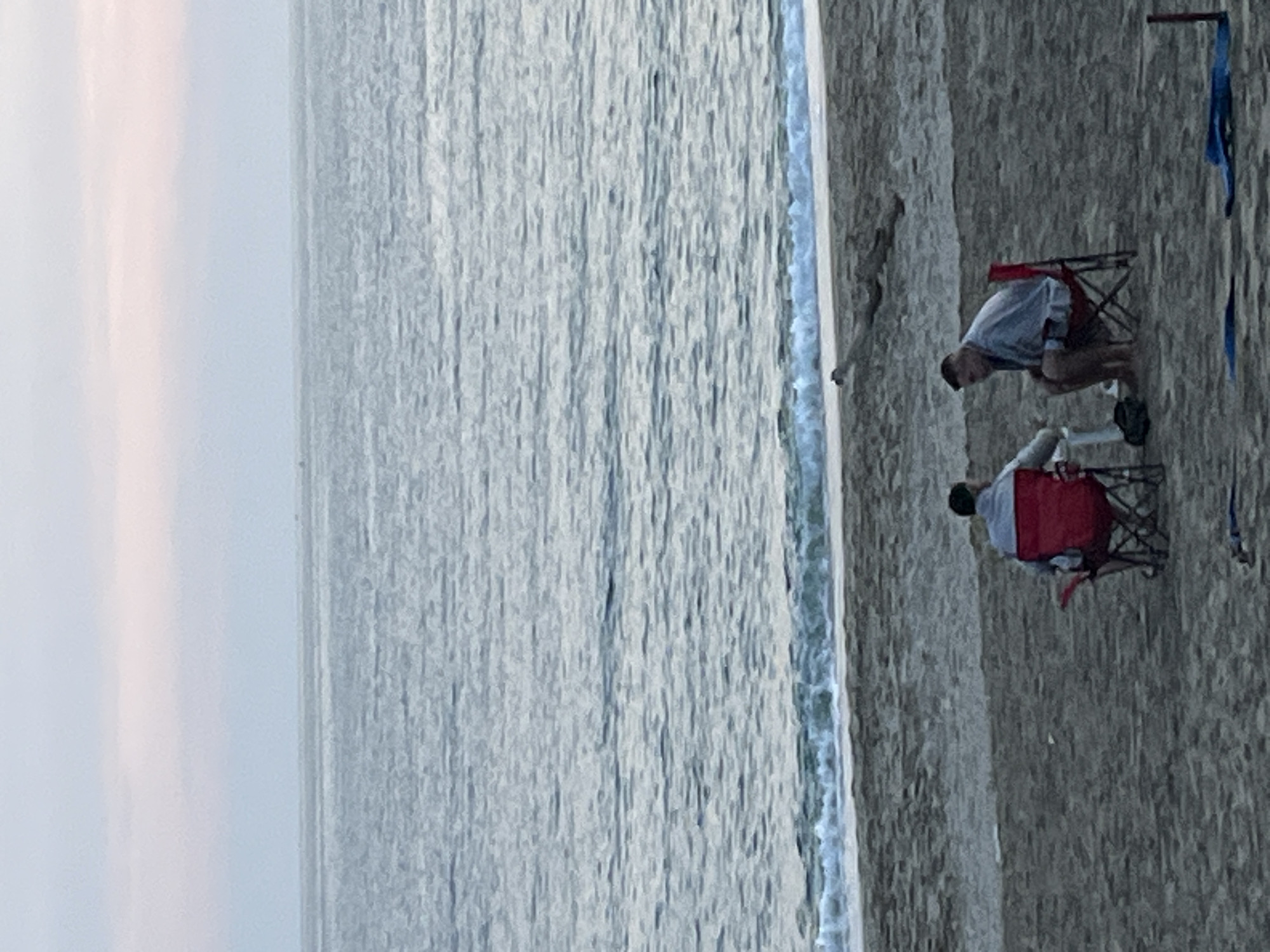 Two people are sitting on beach chairs by the sea. One person is wearing a light-colored shirt and the other is wearing a dark-colored shirt. The sea is to their left and the beach is on the right. The image shows an outdoor scene. Given that the prompt requested the location of Alanya, Turkey, it can be assumed that this is a beach scene in Alanya, Turkey.
