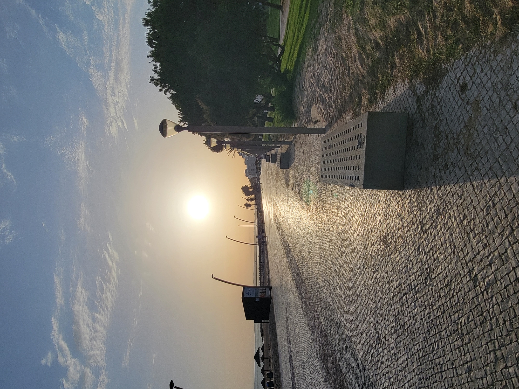 The image depicts a waterfront promenade in Quarteira, Portugal, during what appears to be sunrise or sunset. The sun is visible in the sky, casting a warm glow across the scene. A paved walkway, made of interlocking stones, stretches into the distance, lined with streetlights and benches. To the left of the walkway is a sandy beach with umbrellas, and beyond that, the calm sea. To the right, there is a grassy area with trees, some of which are pine trees. A few people are visible walking along the promenade in the distance. The overall atmosphere is serene and peaceful.