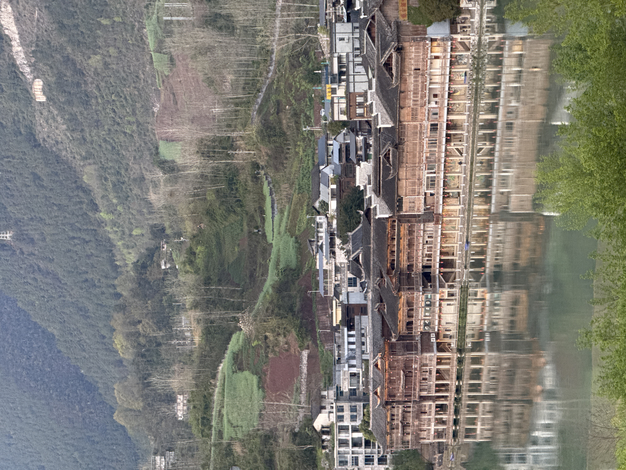 An aerial shot looking downward depicts the city of Zhuoshui, China, with mountain slopes on the left. Zhuoshui’s infrastructure includes homes and buildings along the slopes, and a river on the right with building reflections. Trees on the right edge of the river are also visible.