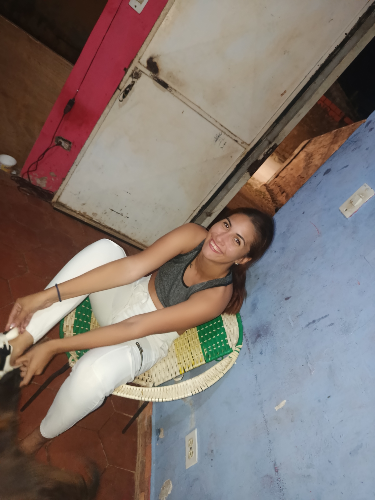 A young woman is seated on a low, woven chair in what appears to be an indoor or semi-outdoor setting in Juan Griego, Venezuela. She is smiling directly at the camera, conveying a cheerful expression. She wears a grey sports bra or crop top and white pants or leggings. Her dark hair is pulled back.
The chair she is sitting on features a green and natural-colored woven pattern. Her hands are positioned near her feet, possibly adjusting something or resting.
The background consists of various textured surfaces. To the left, a reddish-pink wall or panel is visible, with an electrical cord plugged into an outlet. Above and behind her, a large, possibly metal, off-white door or panel shows signs of wear and age. To the right of the woman, a blue-painted wall features another electrical outlet and a light switch. The floor beneath her is covered with terracotta-colored tiles. A blurry patch of brown fur, possibly from a pet, is visible in the bottom left foreground.
The lighting suggests it might be nighttime or an interior space lit by artificial light. The overall environment appears casual and lived-in. No discernible text is visible in the image.