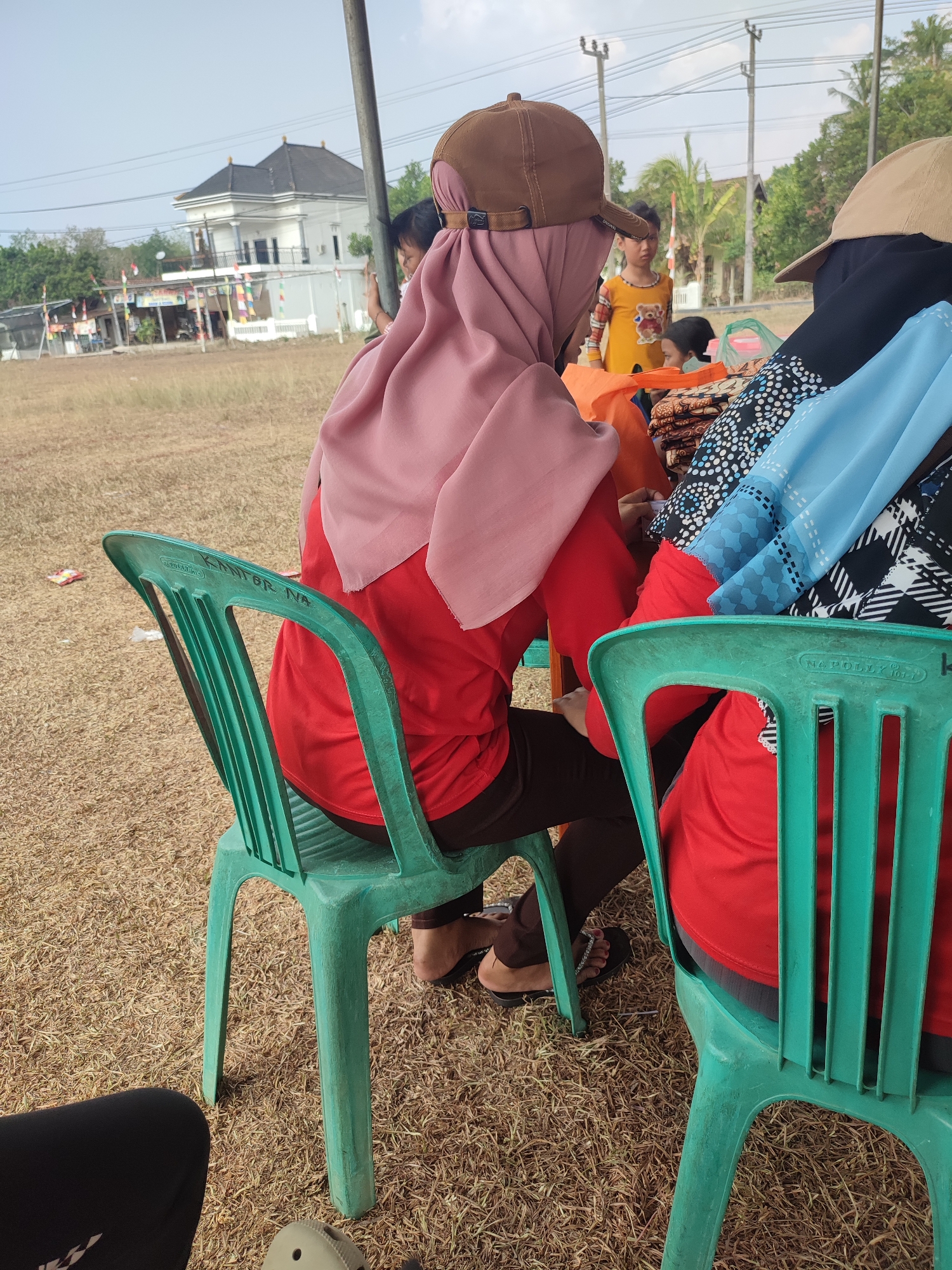 In Nusa Jaya, Indonesia, a medium shot captures an outdoor scene under a bright sky. The foreground features the backs of two individuals seated on green plastic chairs. The person on the left wears a pink hijab and a red shirt, while the person on the right wears a black hijab and a blue and black patterned garment. In the mid-ground, another person in a yellow shirt with a teddy bear graphic stands and looks towards the viewer. Behind them, a two-story building with a dark roof and white walls is visible, along with trees and utility poles. The ground is covered in dry grass. The scene appears to be a casual gathering or event.