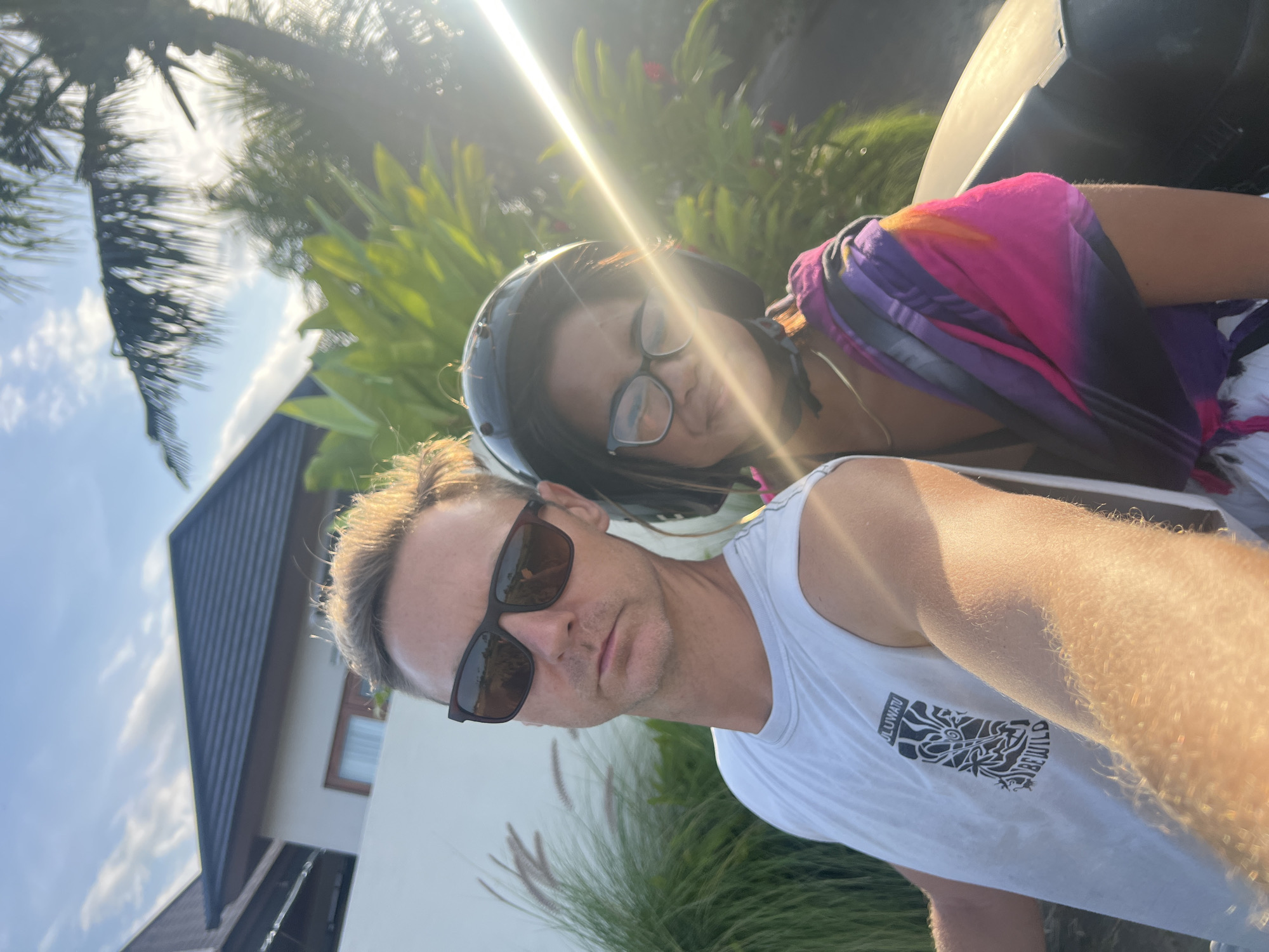 A selfie-style, close-up shot depicts a man and a woman outdoors in the Ubud District, Indonesia. The man, in the foreground, wears sunglasses and a white tank top with a graphic print on it. The woman, positioned behind him, is wearing a helmet and glasses, and a multi-colored garment. The background features a building with a dark gray roof, tropical foliage, and a bright sky with a sun glare.