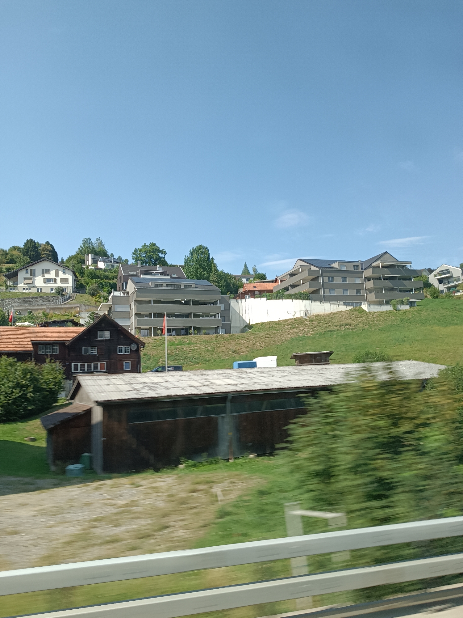 This is an outdoor, daytime, medium-shot photograph taken from a moving vehicle, likely a train or car, as indicated by the motion blur in the foreground and midground. The scene depicts a hillside residential area in Niederteufen, Switzerland, under a clear blue sky with a few wispy clouds.
In the foreground, there is a white metal guardrail, with green foliage and a gravel path visible behind it. A portion of a low, wooden shed-like structure with a corrugated metal roof is also present in the immediate foreground.
Dominating the midground and background are several houses and apartment buildings situated on a grassy slope. In the lower-midground, a dark wooden house with a red-tiled roof features several windows. To its right, a modern, multi-story apartment building with a grey facade, balconies, and solar panels on the roof is prominent. Two red flags are visible on flagpoles in front of this building. Further up the hill, a lighter colored, multi-story house is visible. In the upper left, another white house with brown shutters sits on a terraced slope. The hillside is covered in lush green grass and mature trees.
The overall impression is of a tranquil, suburban or semi-rural setting in Switzerland. There are no people visible in the image.