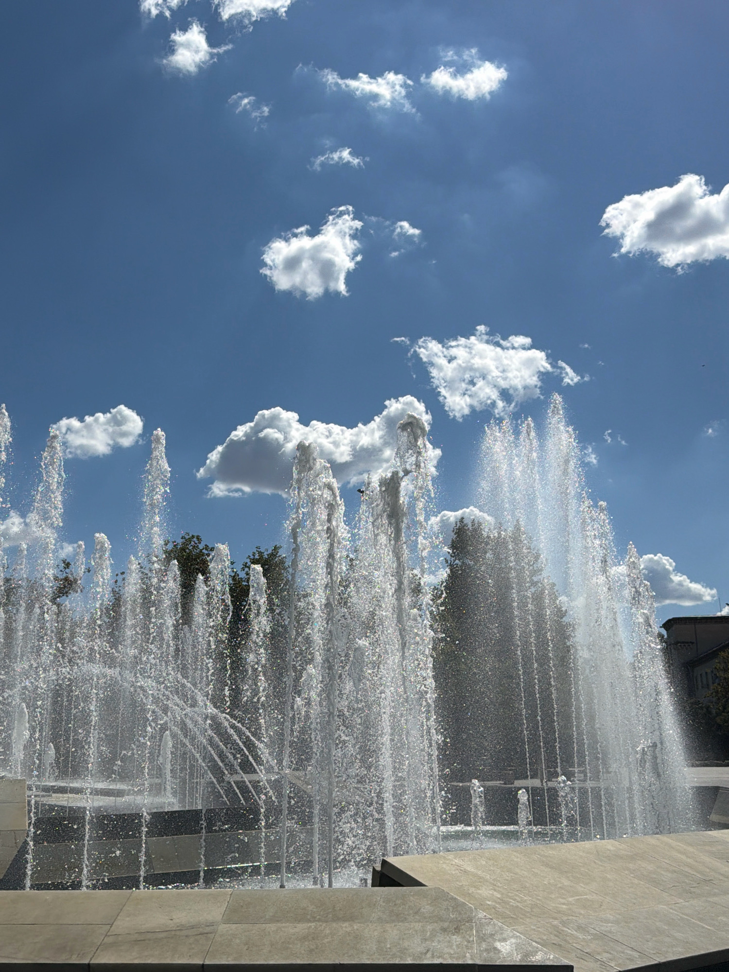 The image captures a dynamic water fountain on a bright, sunny day. The sky above is a vibrant blue, dotted with fluffy white clouds. The fountain's water sprays upwards in multiple streams, creating a shimmering, translucent curtain. The water is catching the sunlight, causing it to sparkle. In the background, the tops of green trees are visible, and a portion of a building can be seen on the right side. The foreground consists of concrete slabs and tiered structures that form the base of the fountain area. The scene is devoid of any people. The fountain is located in Pleven, Bulgaria, at the Kaylaka Park, specifically near the central fountain complex.